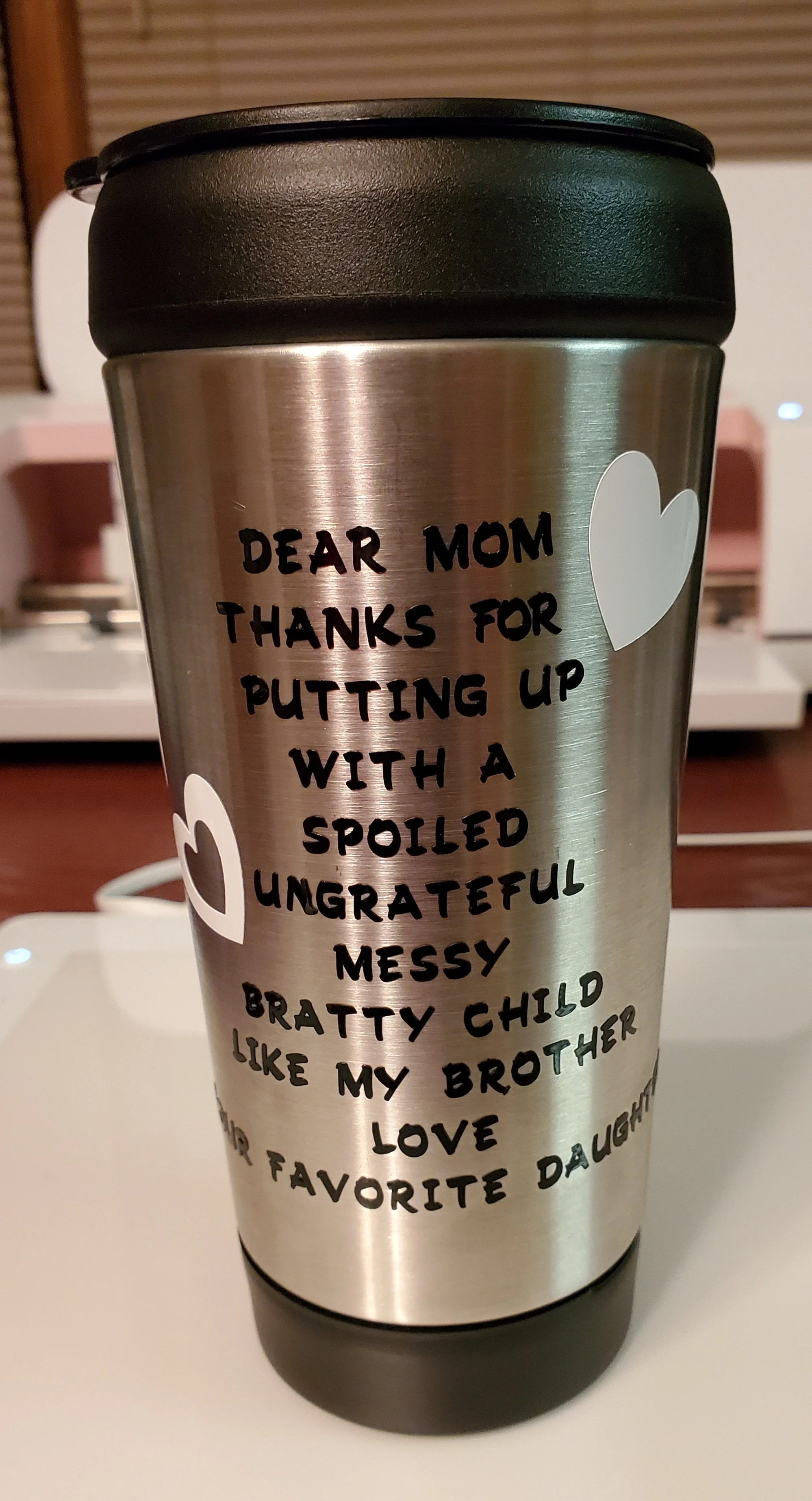 A stainless steel travel mug with black lid, decorated with white heart stickers and a heartfelt message for a mother.