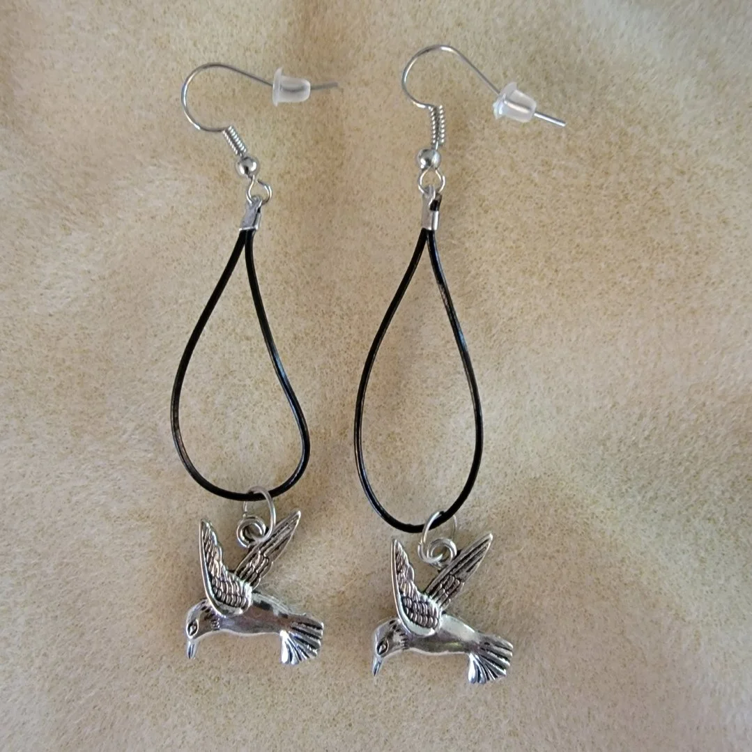 Pair of hummingbird-shaped earrings with black loops and metal hummingbird charms hanging from hooks on a beige background.