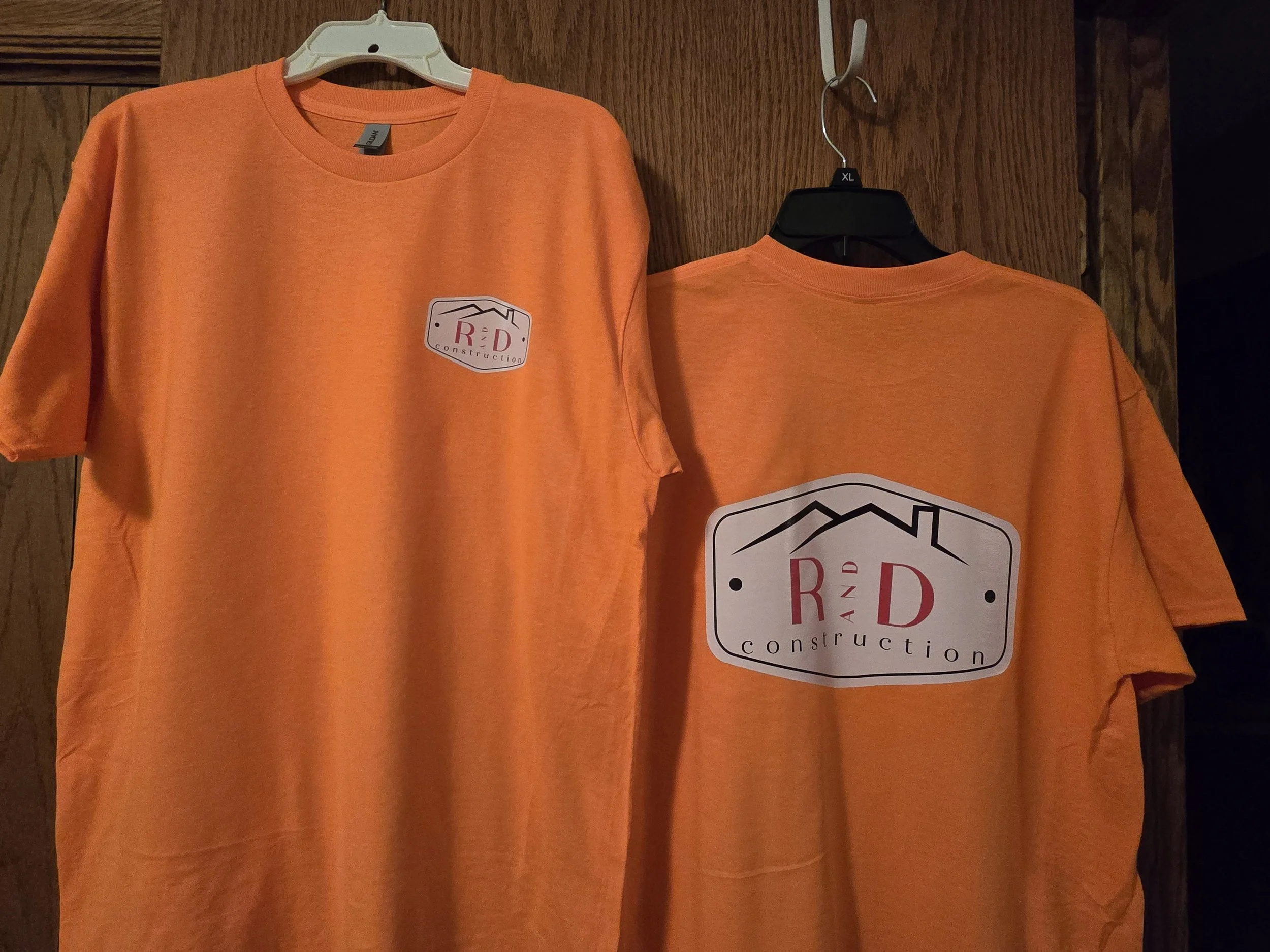 Two orange construction company T-shirts hanging on black and white hangers, one from the front and one from the back, with a logo featuring mountains, a house outline, and the words 'R&D construction'.