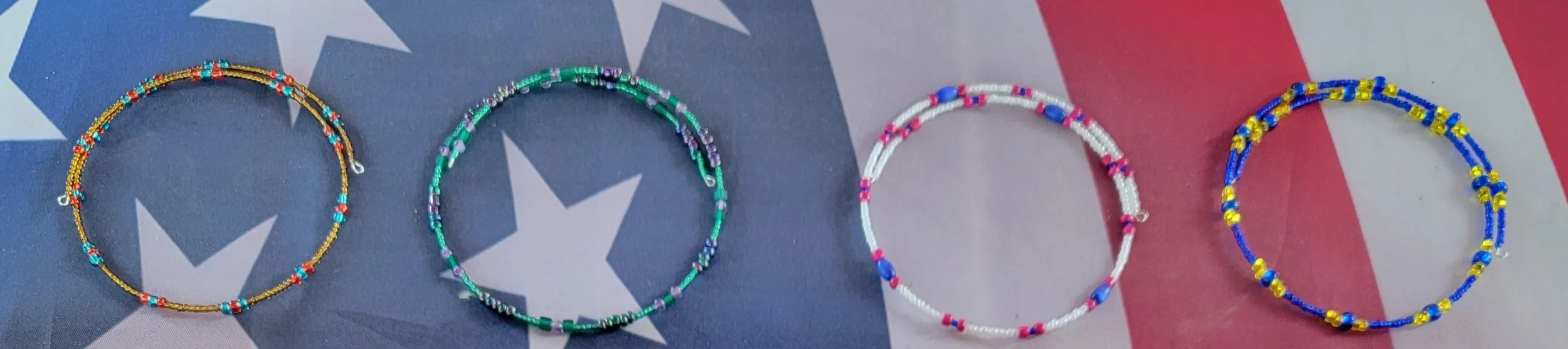 Four colorful beaded necklaces laid out on a fabric background featuring the American flag. The necklaces are in various colors and styles, with the flag's stars and stripes visible behind them.