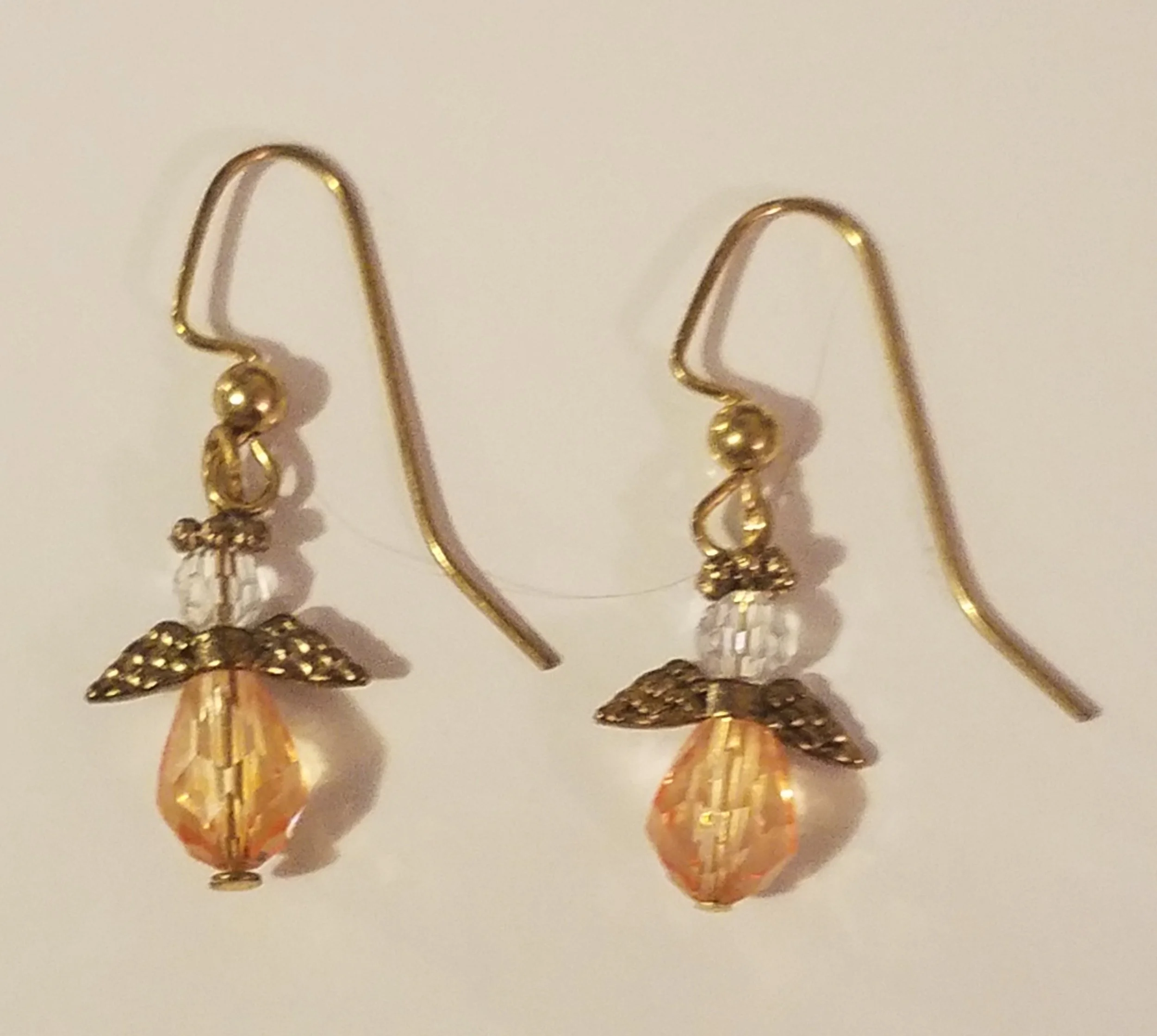 Gold-colored earrings with angel charm, clear bead, peach-colored faceted bead, and leaf accents, hanging on hooks.