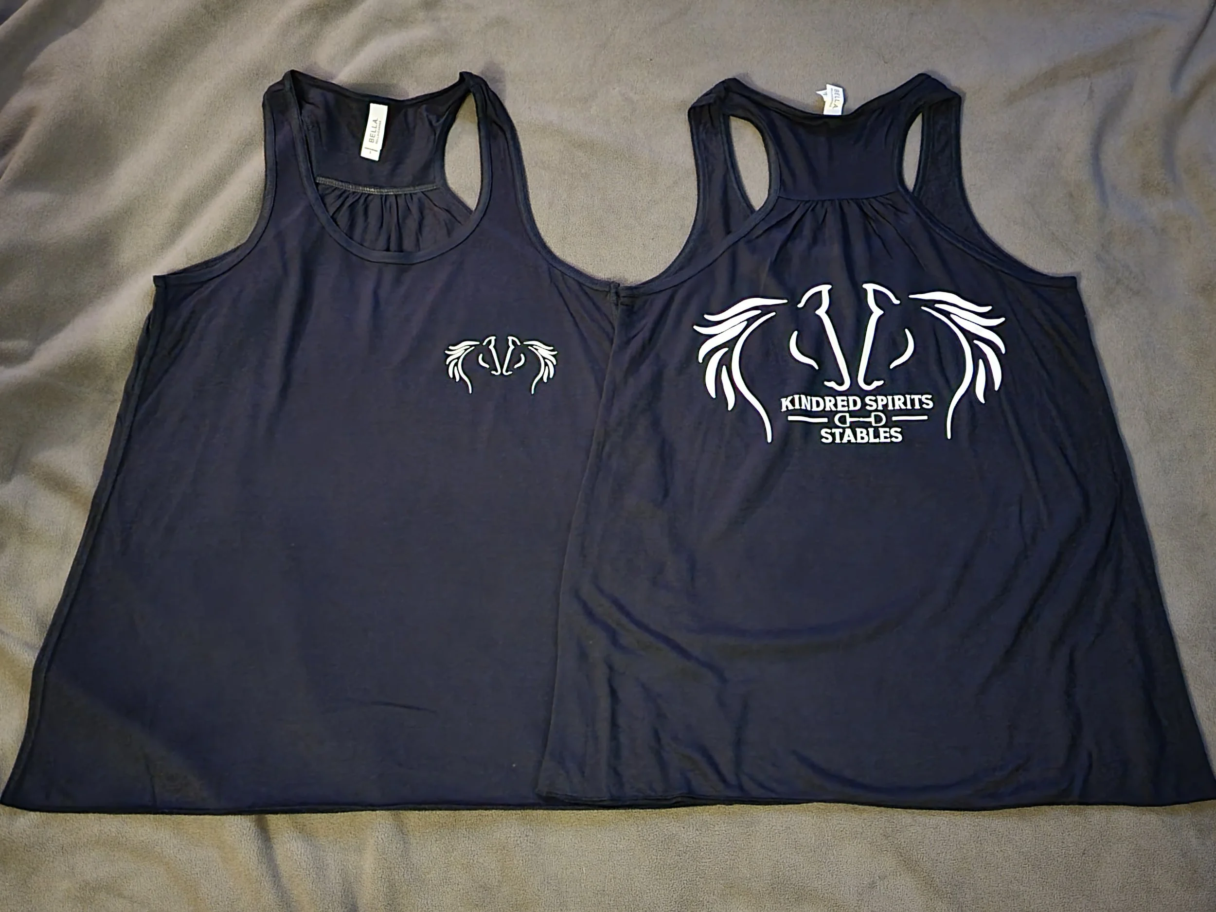 Two navy blue tank tops laid flat on a grey surface, one with a small white logo on the front and the other with a larger logo and text 'Kindred Spirits Stables' on the back.