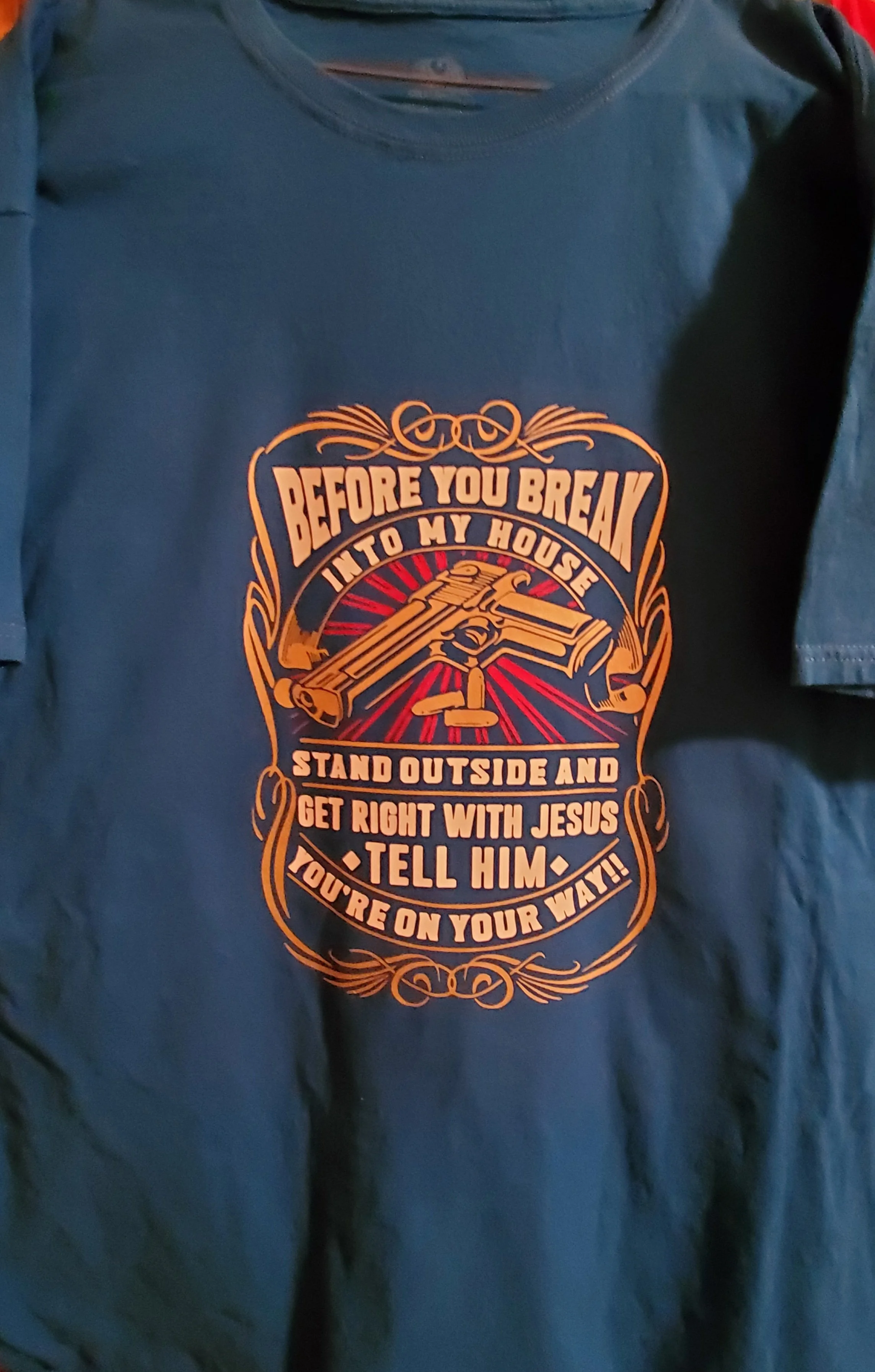 A dark blue T-shirt with a printed design of a gun and text that reads: 'Before you break into my house, stand outside and get right with Jesus. Tell him you're on your way!'
