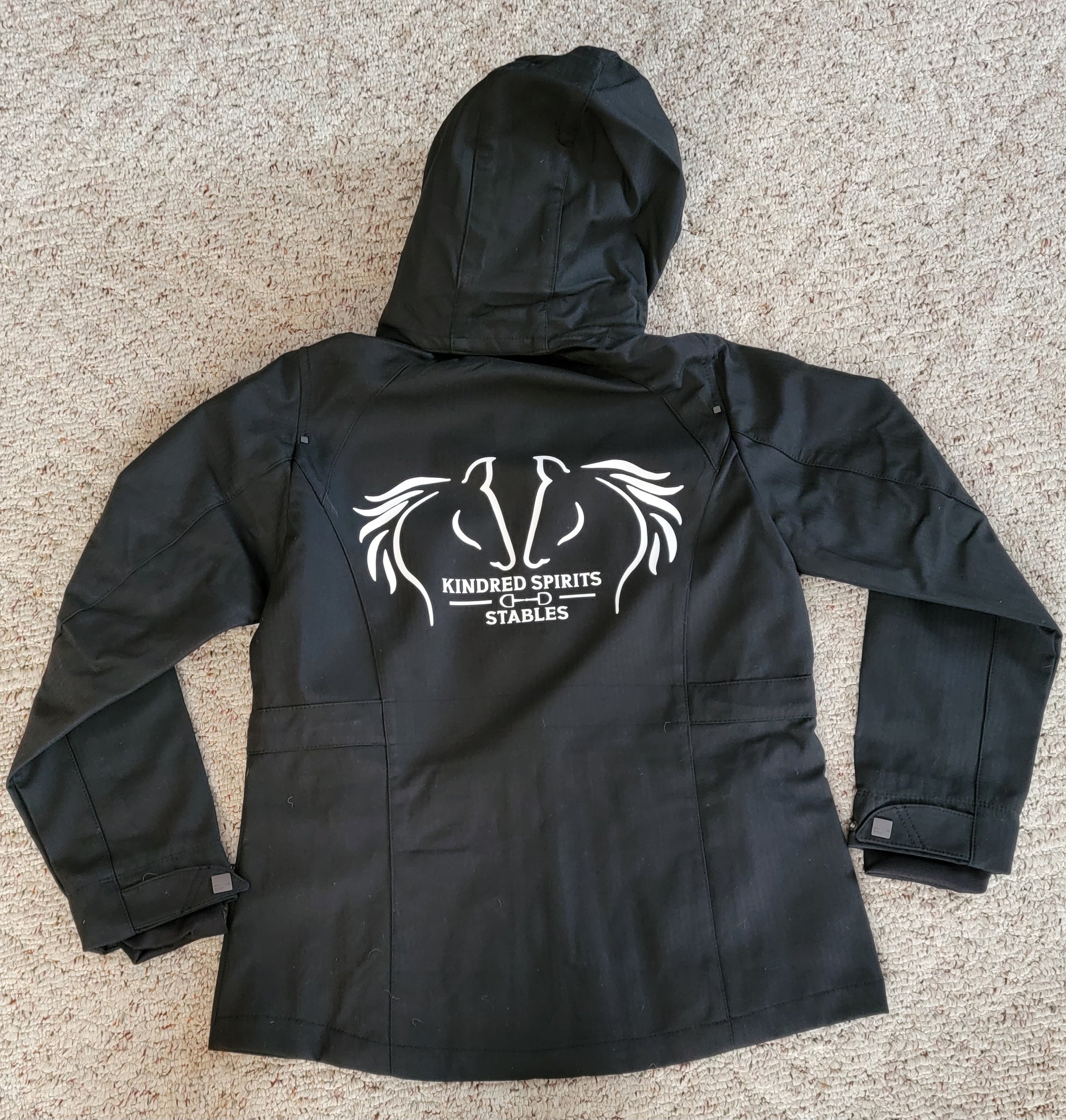 Black jacket with a white logo on the back that says "Kindred Spirits Stables" and features two stylized horse heads facing each other with flowing manes.