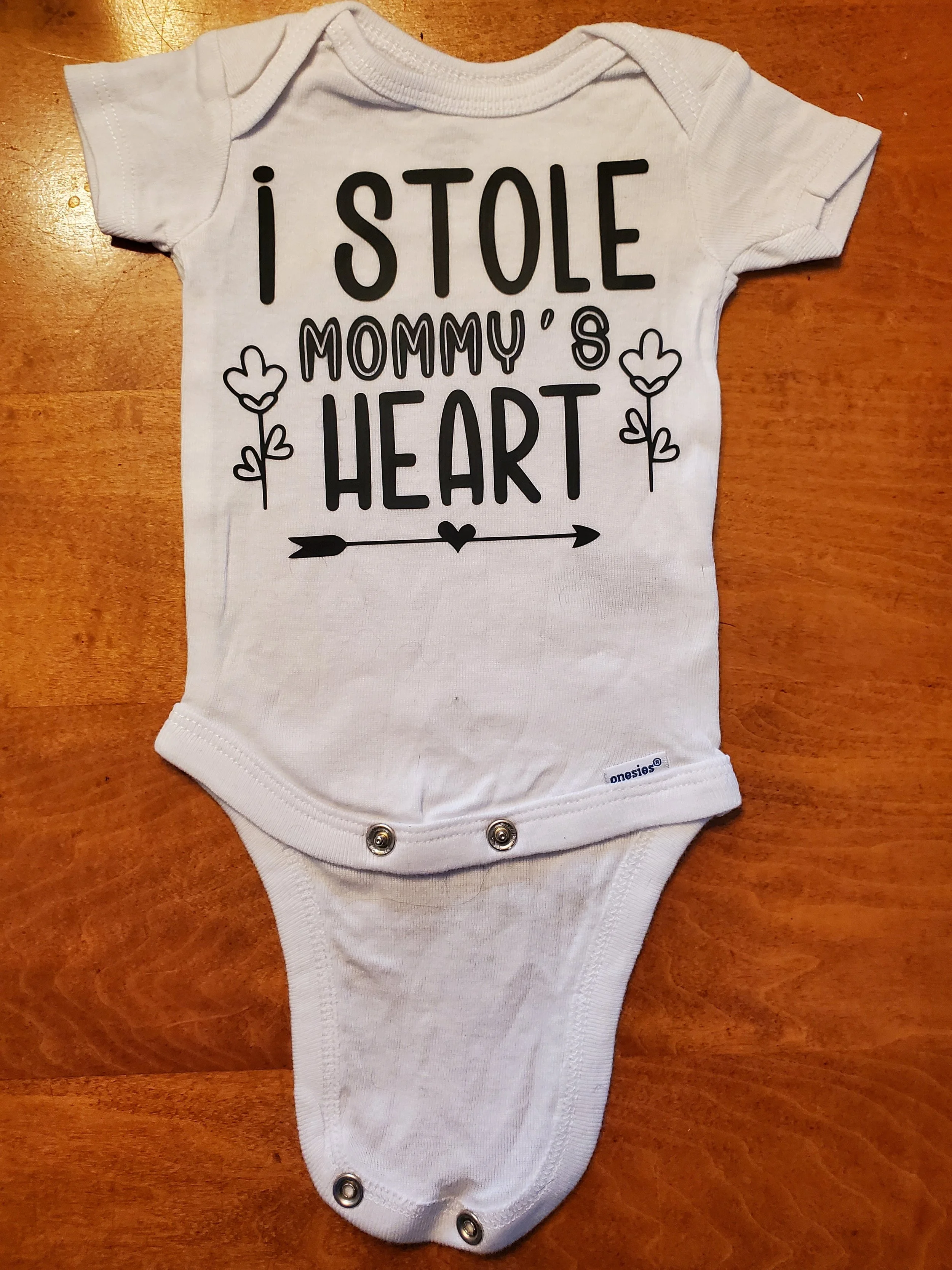 A small white baby onesie with black text that reads, "I stole mommy's heart," decorated with hearts, flowers, and an arrow.
