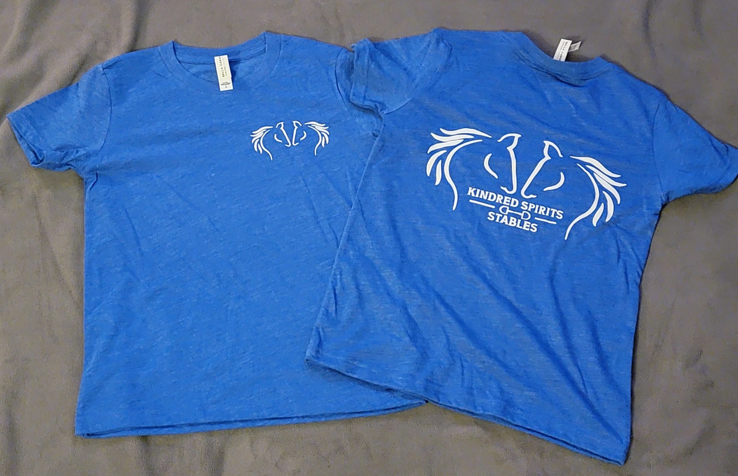 Blue T-shirts, one front and one back, with fantasy-themed horse and wing designs and the text "Kindred Spirits & Stables."