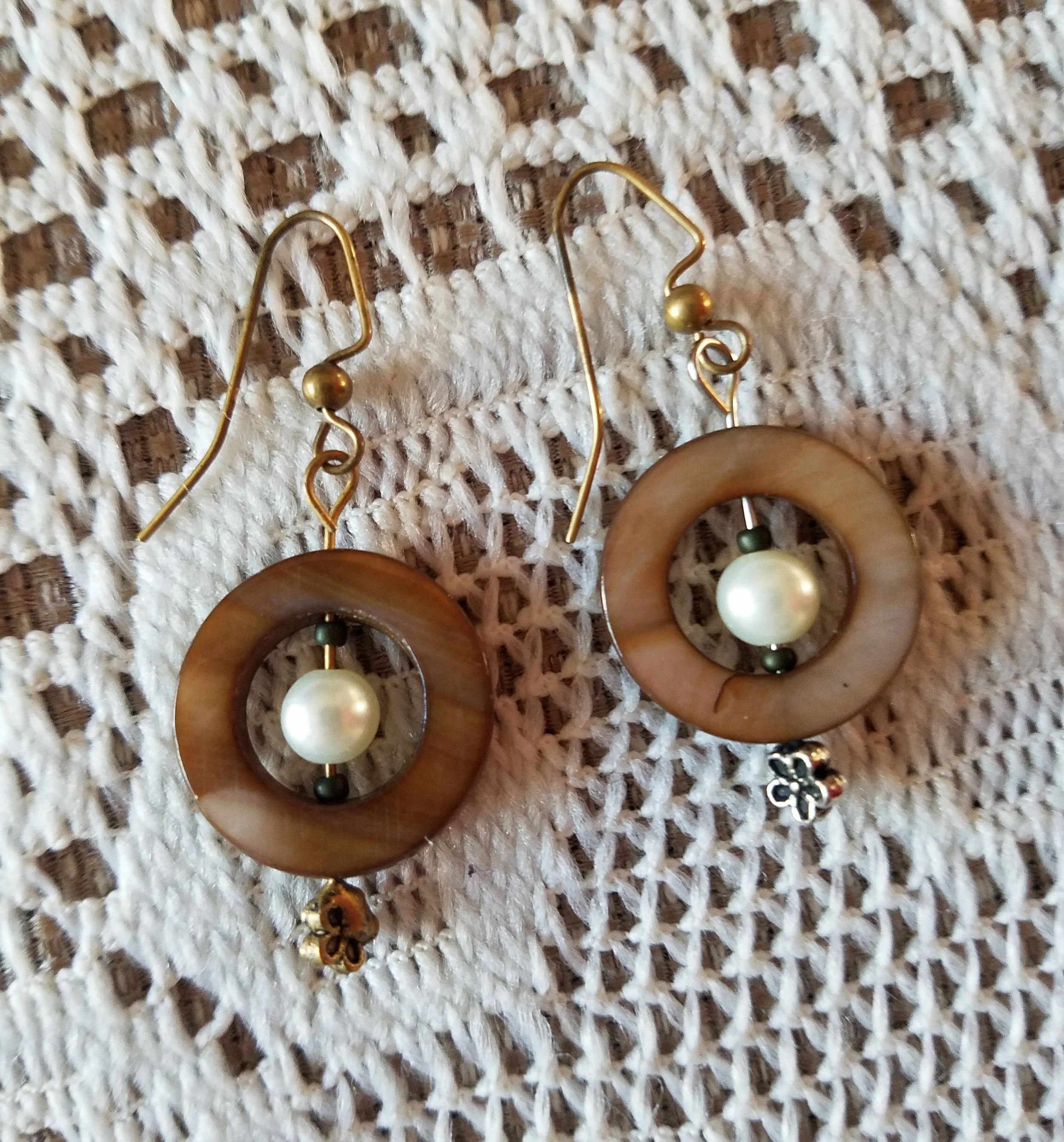 A pair of earrings with circular wooden hoops, pearl beads, and small flower charms, placed on a white woven fabric background.