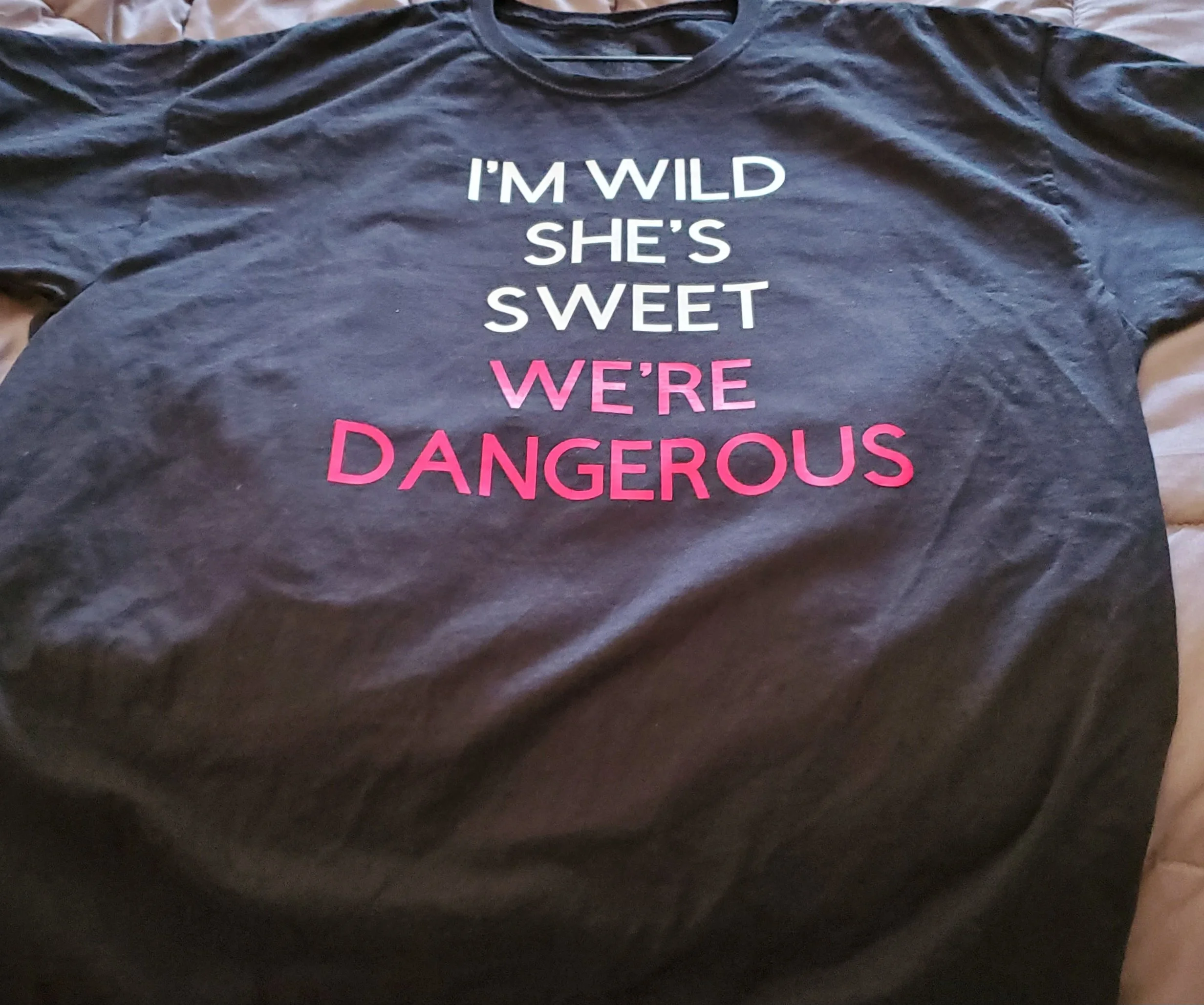 A black T-shirt with the text 'I'M WILD SHE'S SWEET WE'RE DANGEROUS' printed on it, with 'WE'RE DANGEROUS' in pink and the rest in white.