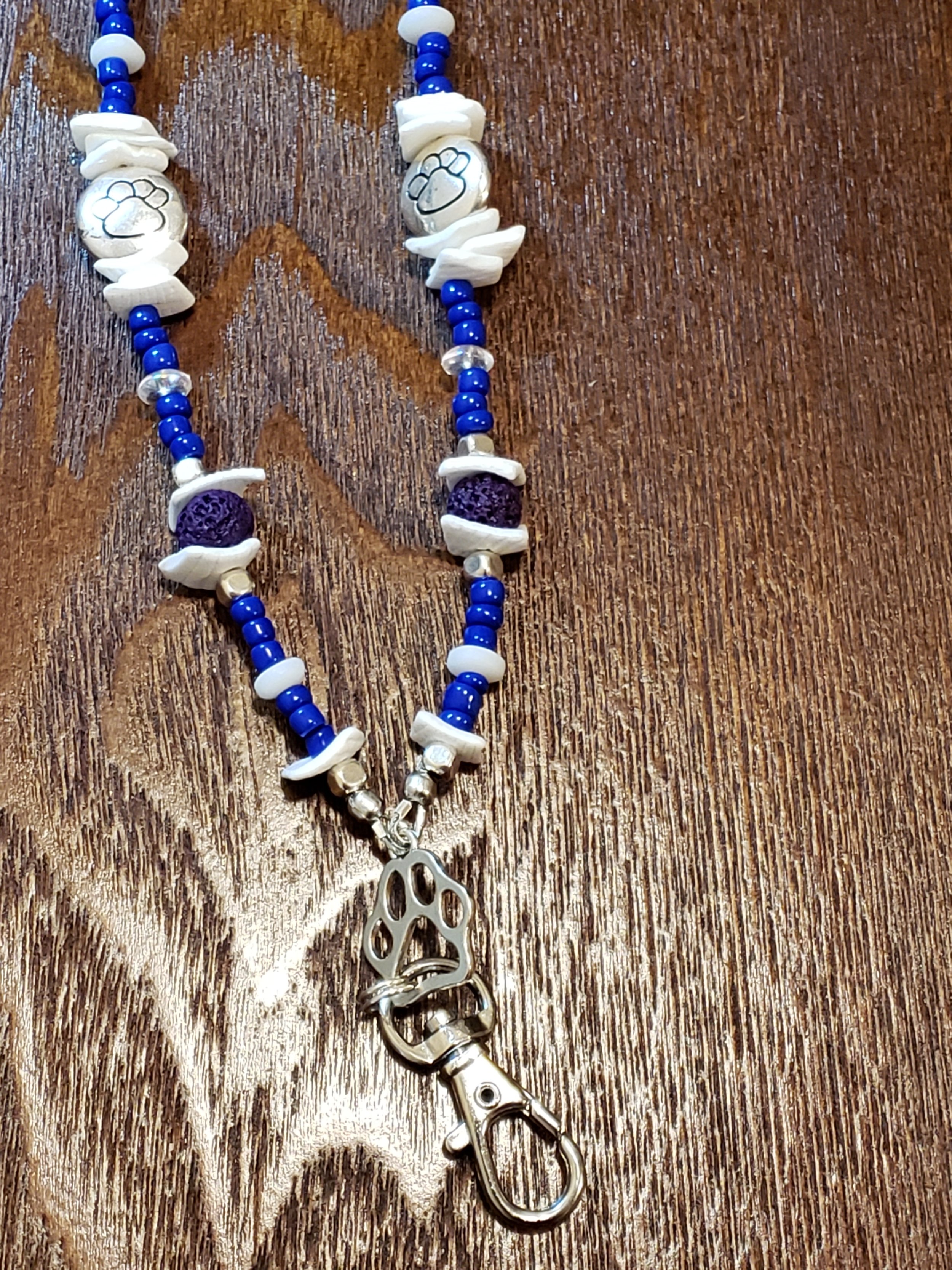 A beaded necklace with white, blue, and purple beads, featuring a silver paw-shaped charm with a key clip at the end, on a wooden surface.