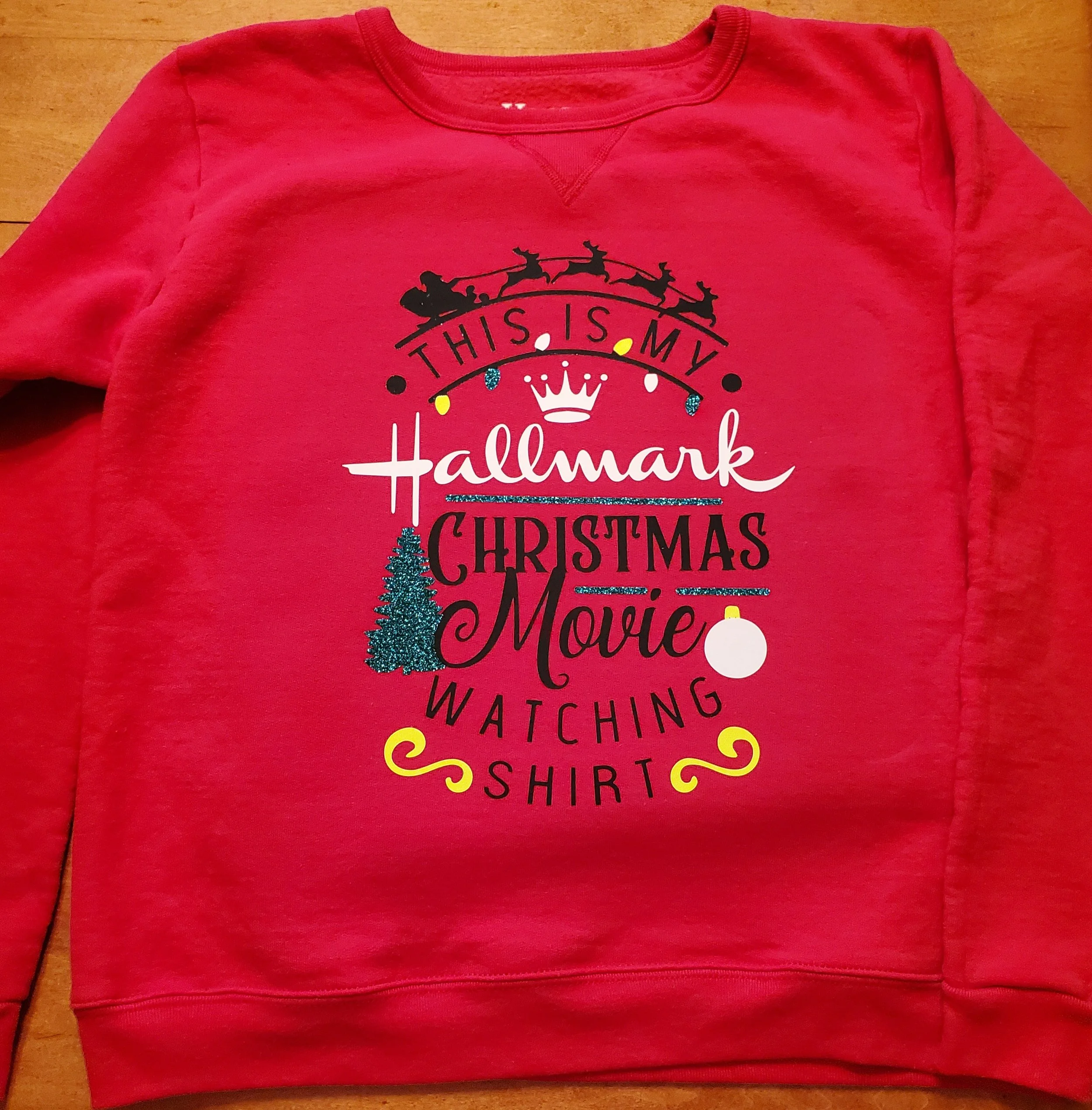 Red sweatshirt with holiday-themed text and graphics, including a Santa sleigh, a Christmas tree, a Christmas ornament, and decorative yellow swirls.
