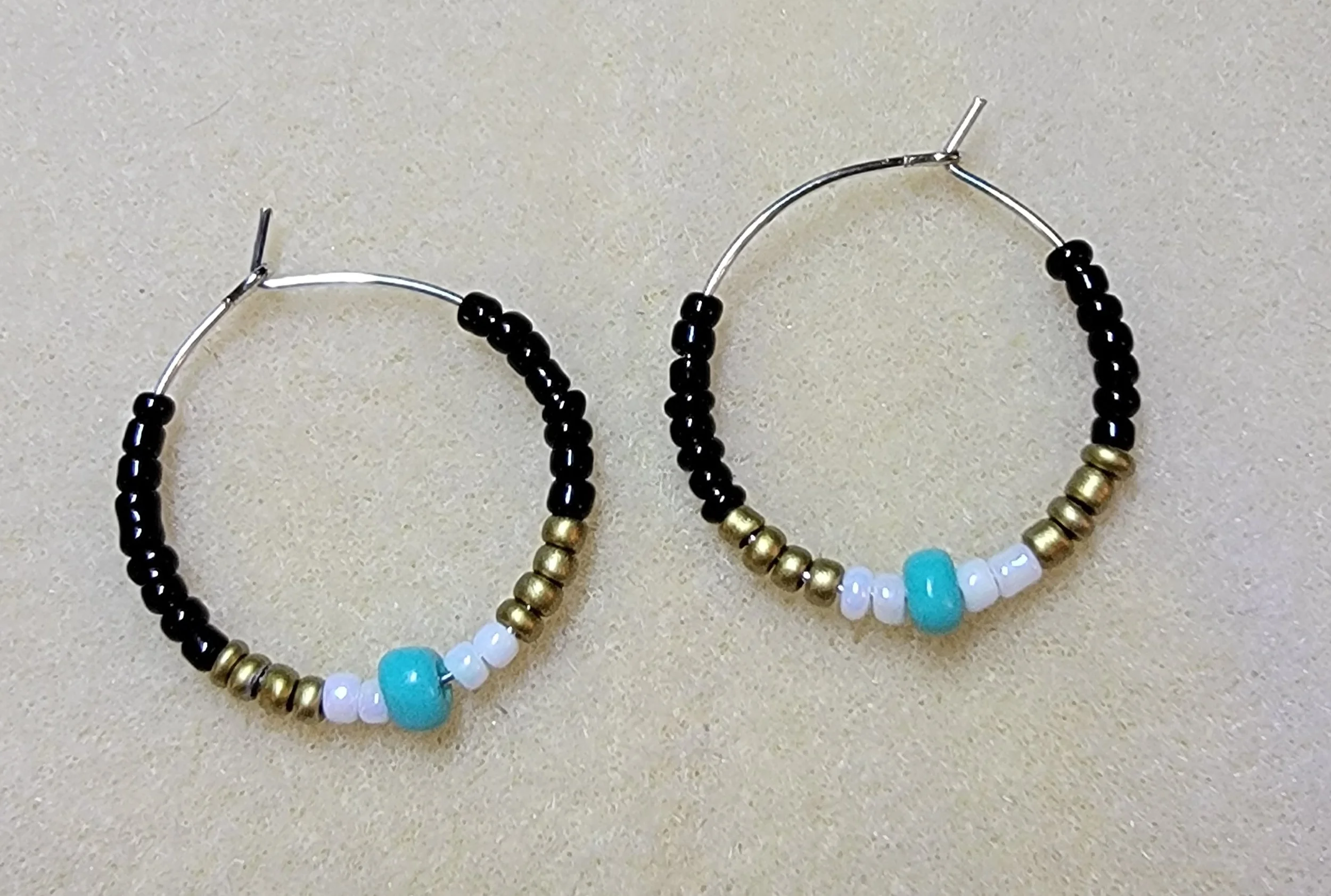 Pair of beaded hoop earrings with black, gold, white, and blue beads, on a beige background.
