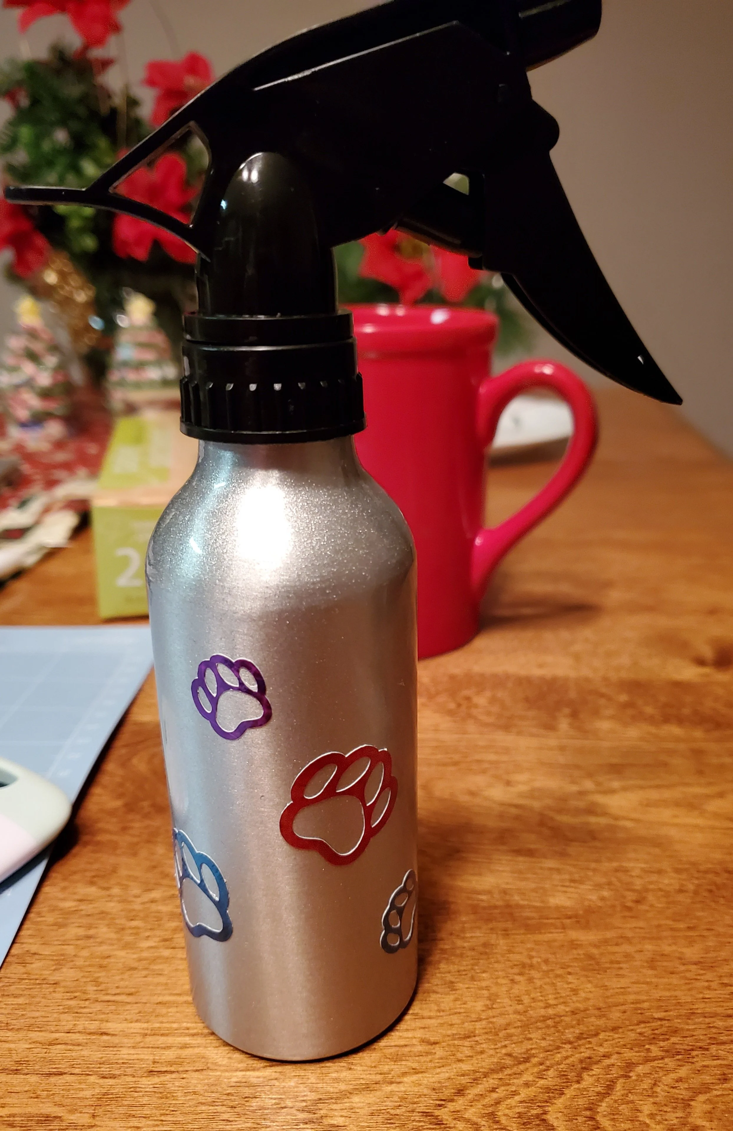 A silver spray bottle with colorful paw print stickers on a wooden table, with a red mug and holiday decorations in the background.