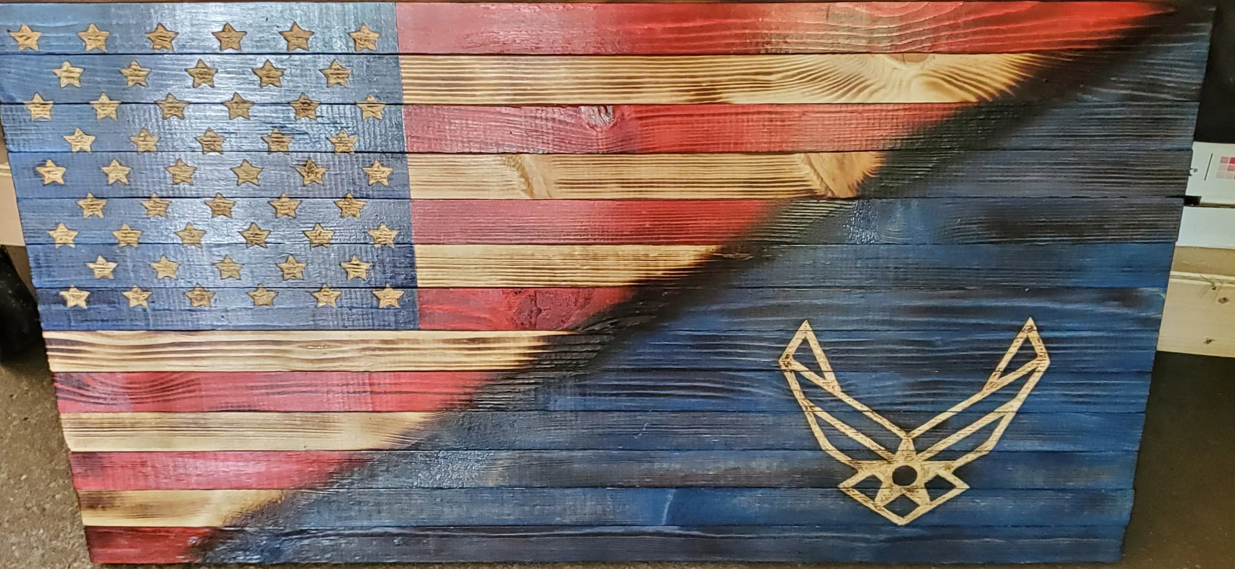 A wooden American flag with red, white, and blue painted stripes and gold star shapes; there is a gold outlined wings of freedom symbol on the lower right side.