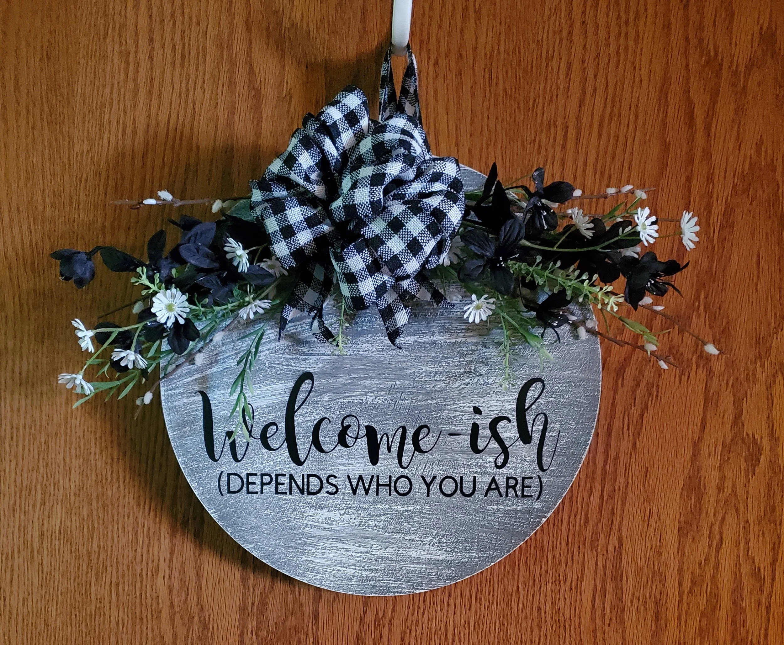 A decorative wall hanging with the words "welcome-ish (depends who you are)" written on a round, silver-finish wooden sign. The sign is adorned with faux flowers and a black-and-white gingham bow.