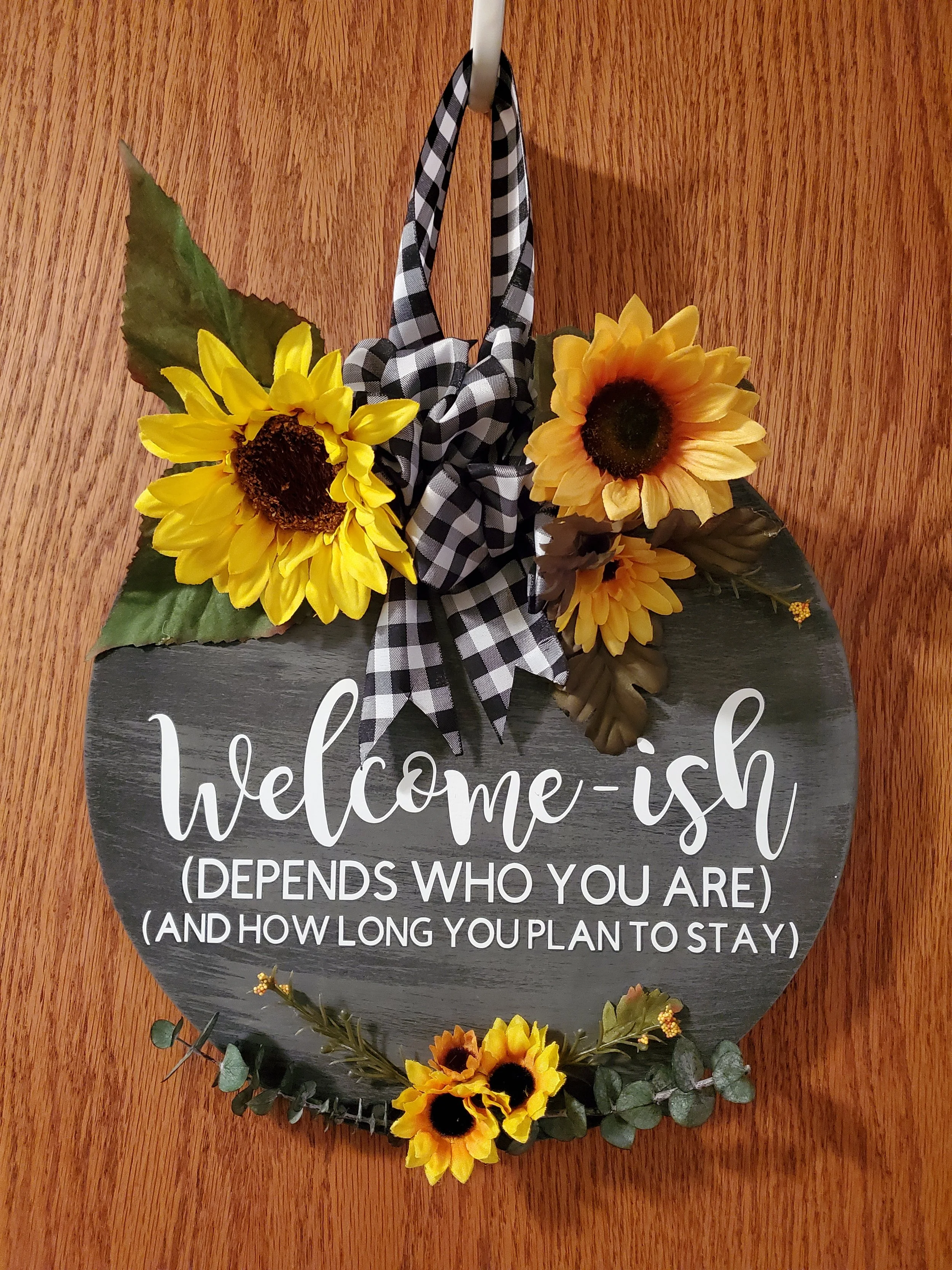 Decorative round sign with sunflowers, eucalyptus leaves, a black and white checkered bow, and the quote 'welcome-ish (depends who you are) (and how long you plan to stay)'