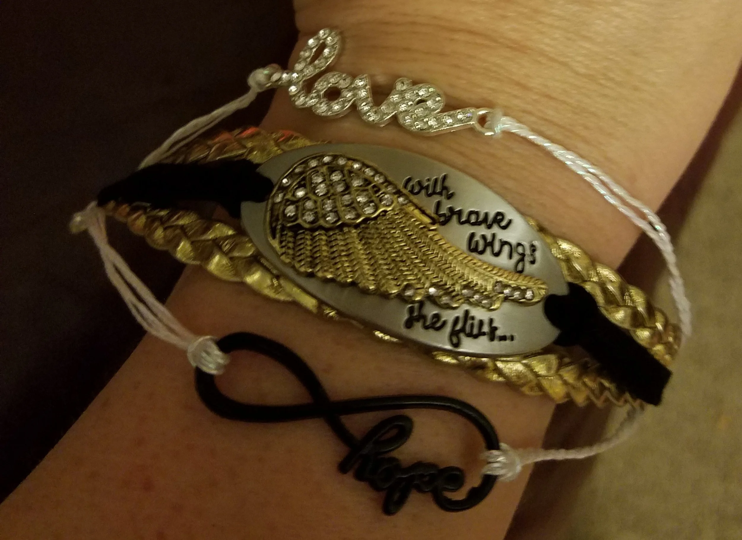 Close-up of a wrist wearing multiple bracelets, including a silver bracelet with the word 'love', a braided gold-colored bracelet, a black hair tie with a wire word 'hope', and a prominent bracelet with a sea shell and the words 'You are the first...