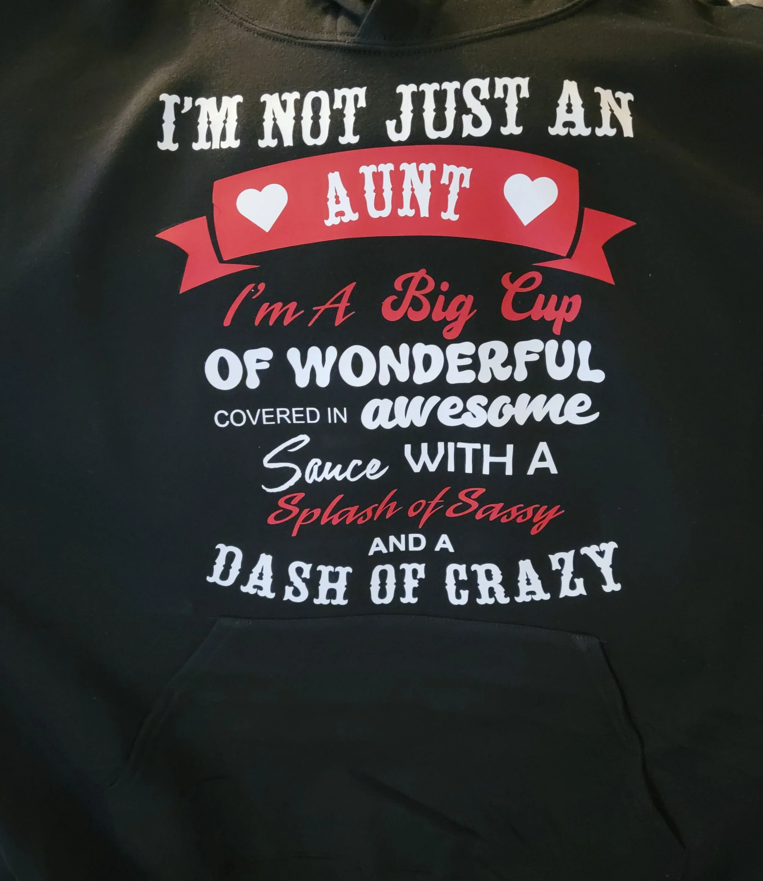 Black hoodie with humorous text about being an aunt, mentioning being a big cup of wonderful, awesome sauce, and a dash of crazy, with decorative hearts and banner.