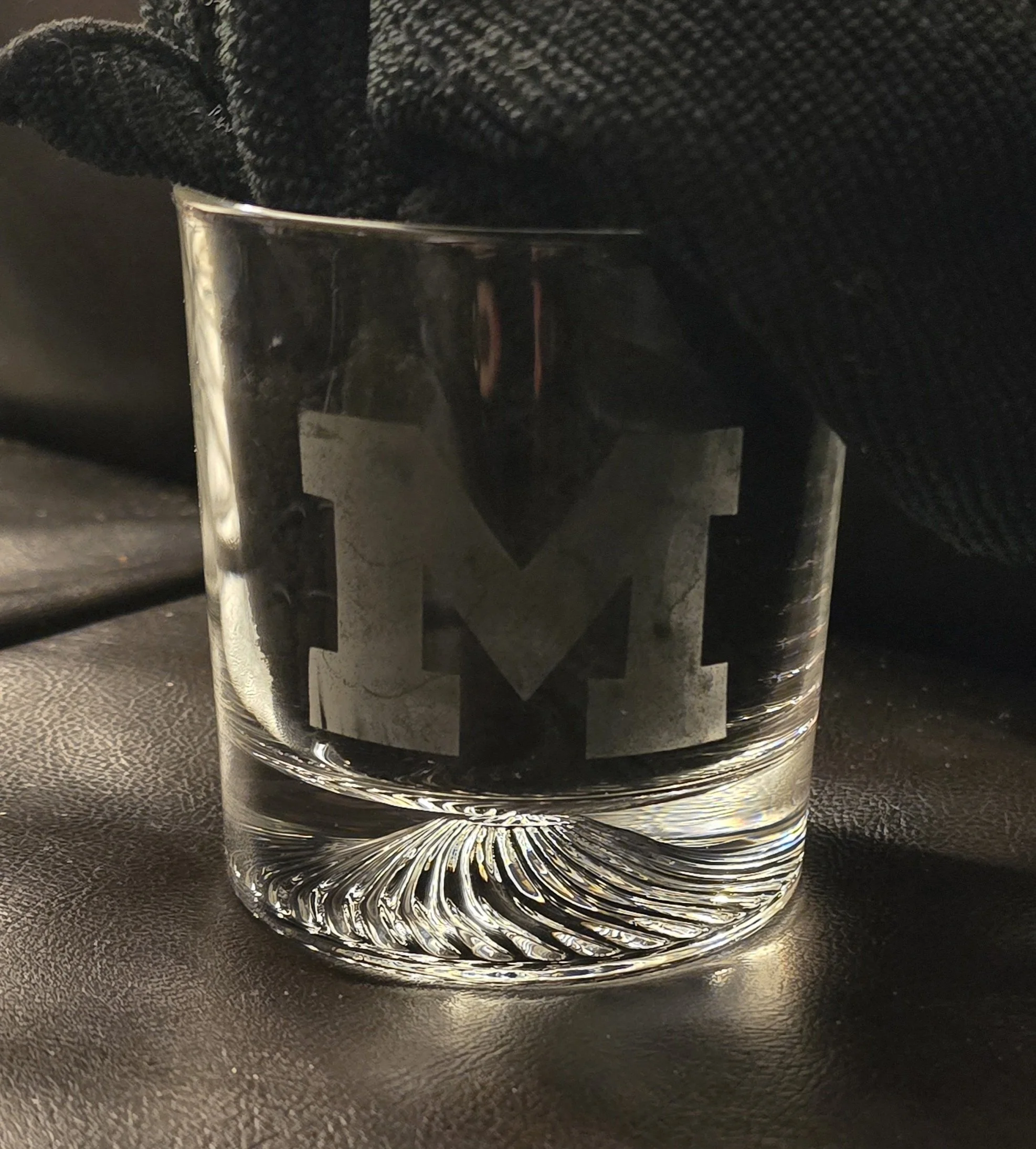 Clear glass with a black logo, filled with a transparent liquid. The glass is on a textured dark surface with a black fabric in the background.