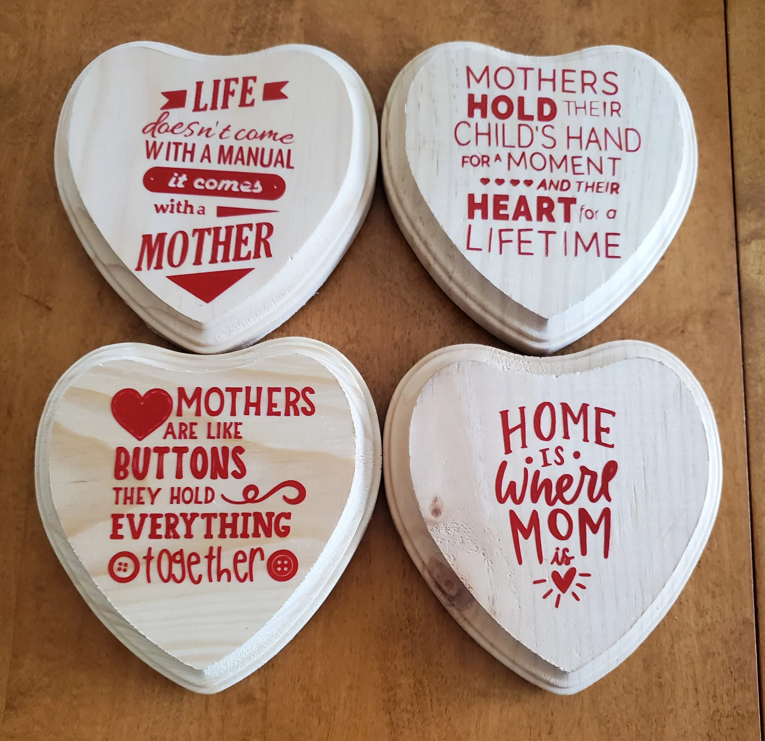 Four heart-shaped wooden plaques with red text and designs, featuring messages about mothers and home, placed on a wooden surface.