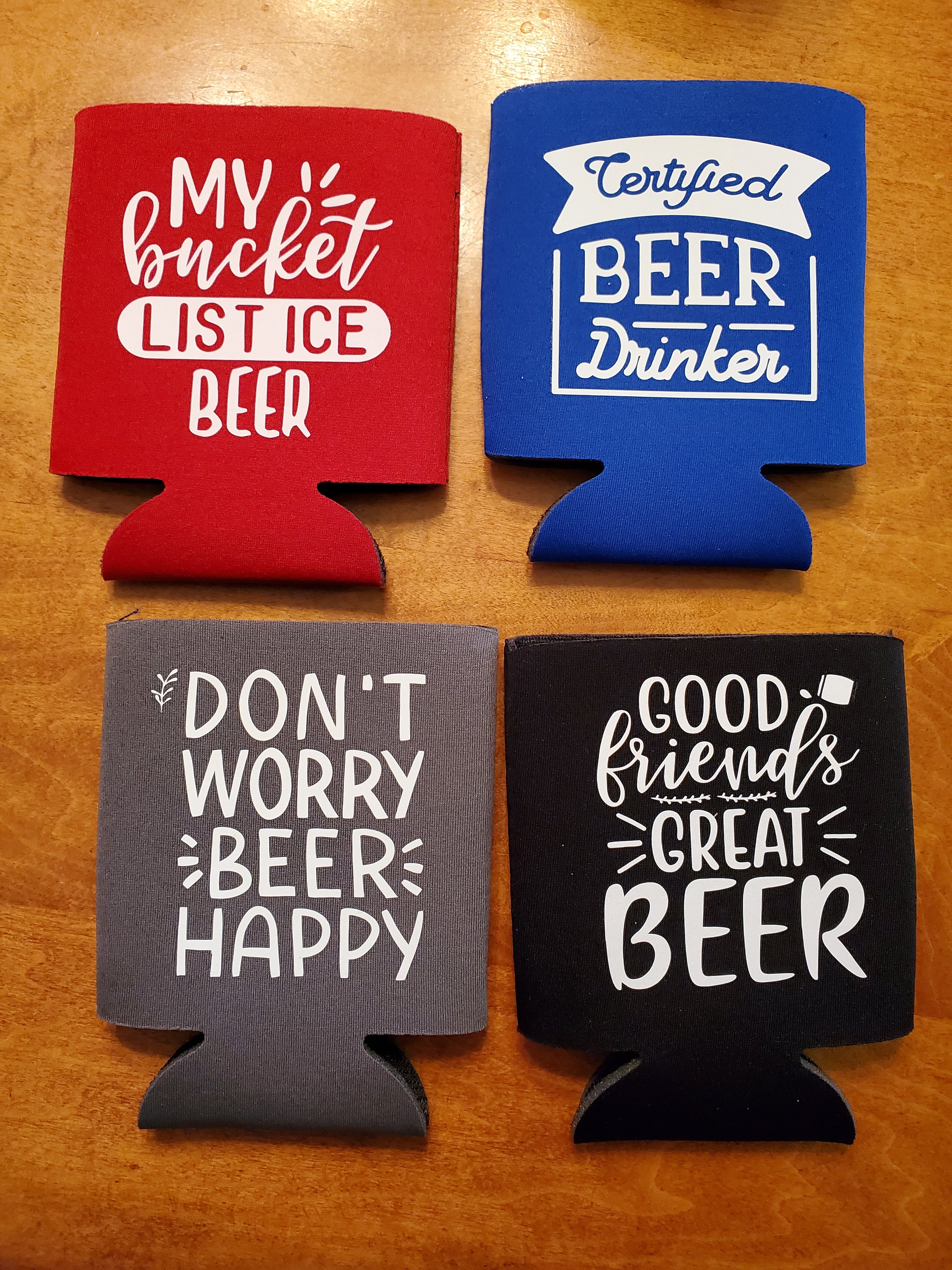 Four colorful drink koozies with beer-related quotes on a wooden surface.