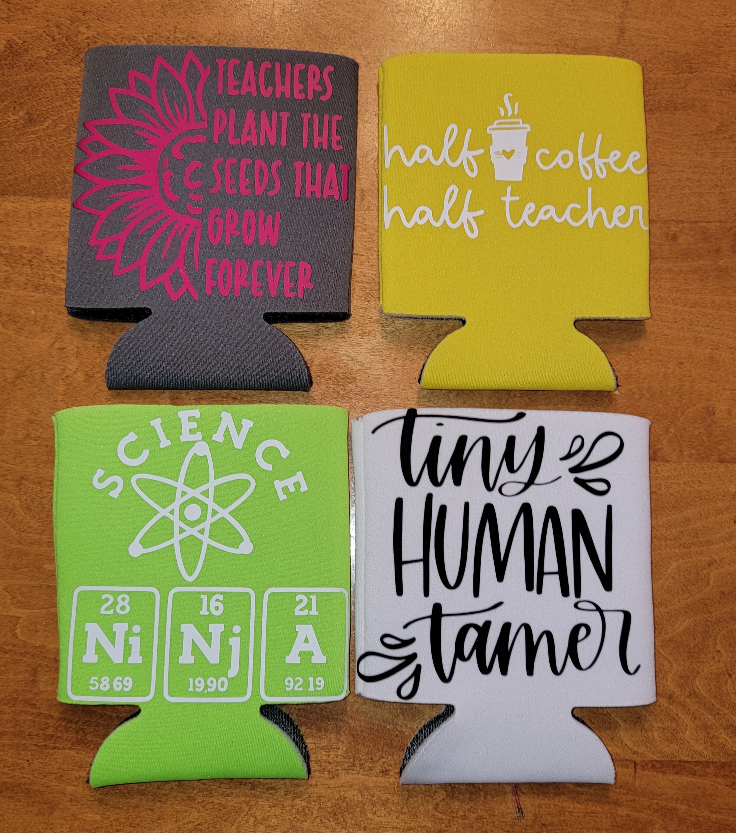 Four drink koozies with different designs and messages, placed on a wooden surface.