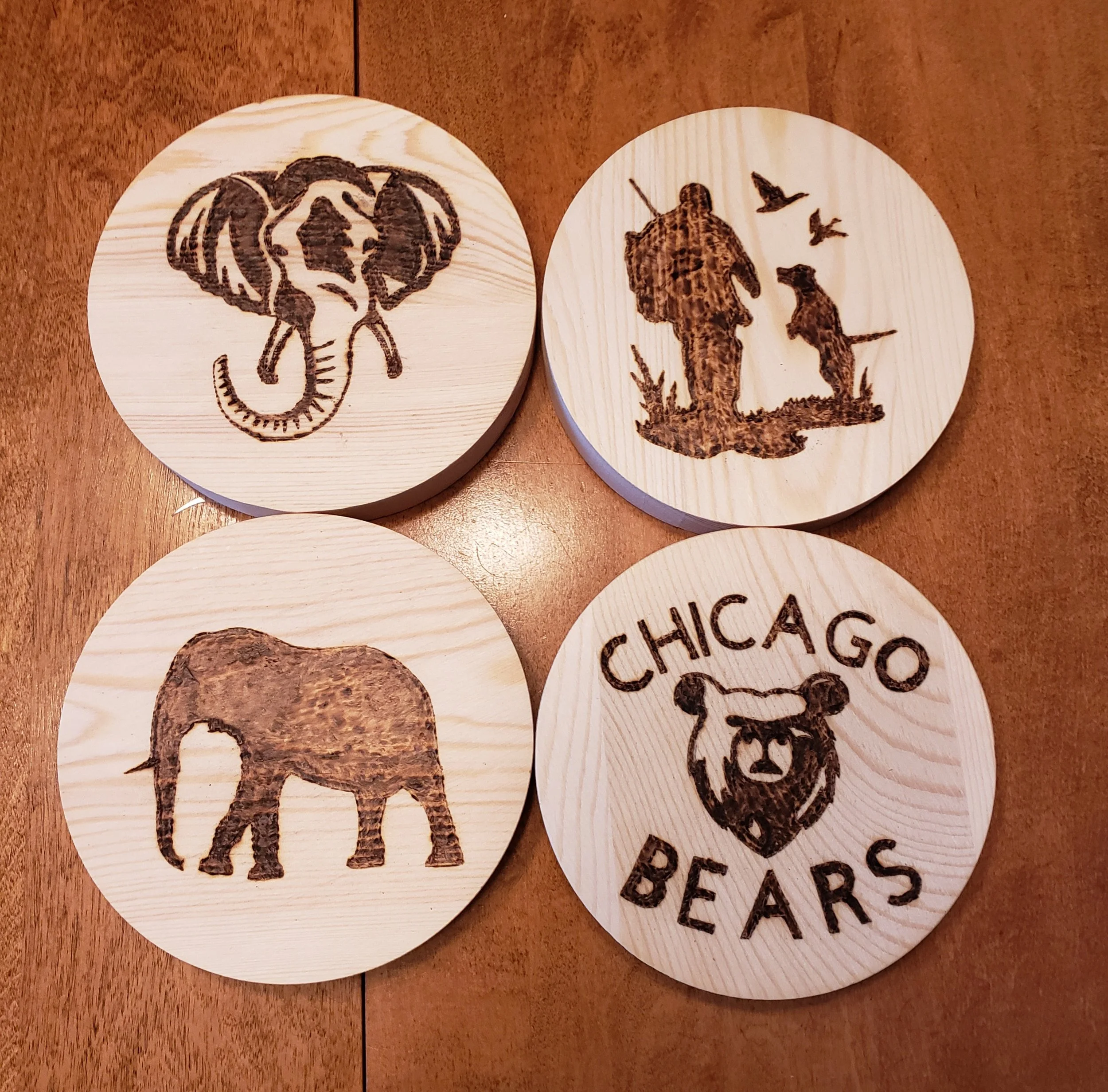 Four wooden coasters with burnt wood designs: an elephant, a hunter with a dog and birds, an elephant, and the Chicago Bears logo with a bear's face and text.