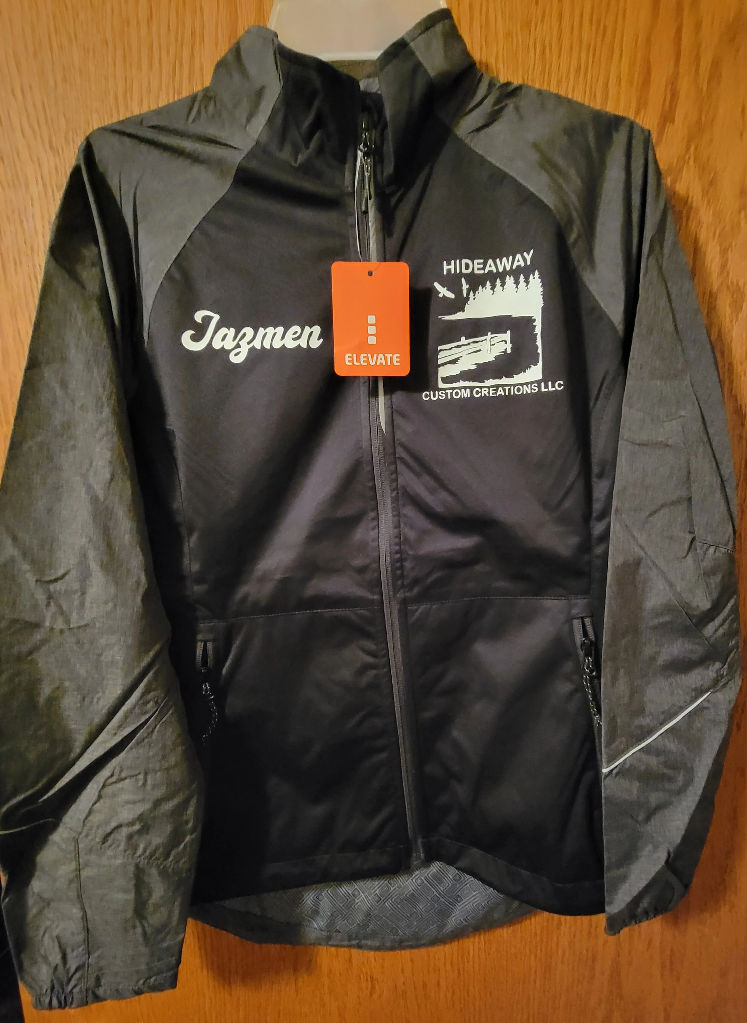 A black and gray jacket with zippered pockets, hanging on a wooden surface. The jacket has text and logos on the front, including the names 'Jazmen' and 'HIDEAWAY' with a graphic of trees and a lake. A red tag with the word 'ELEVATE' is attached to t