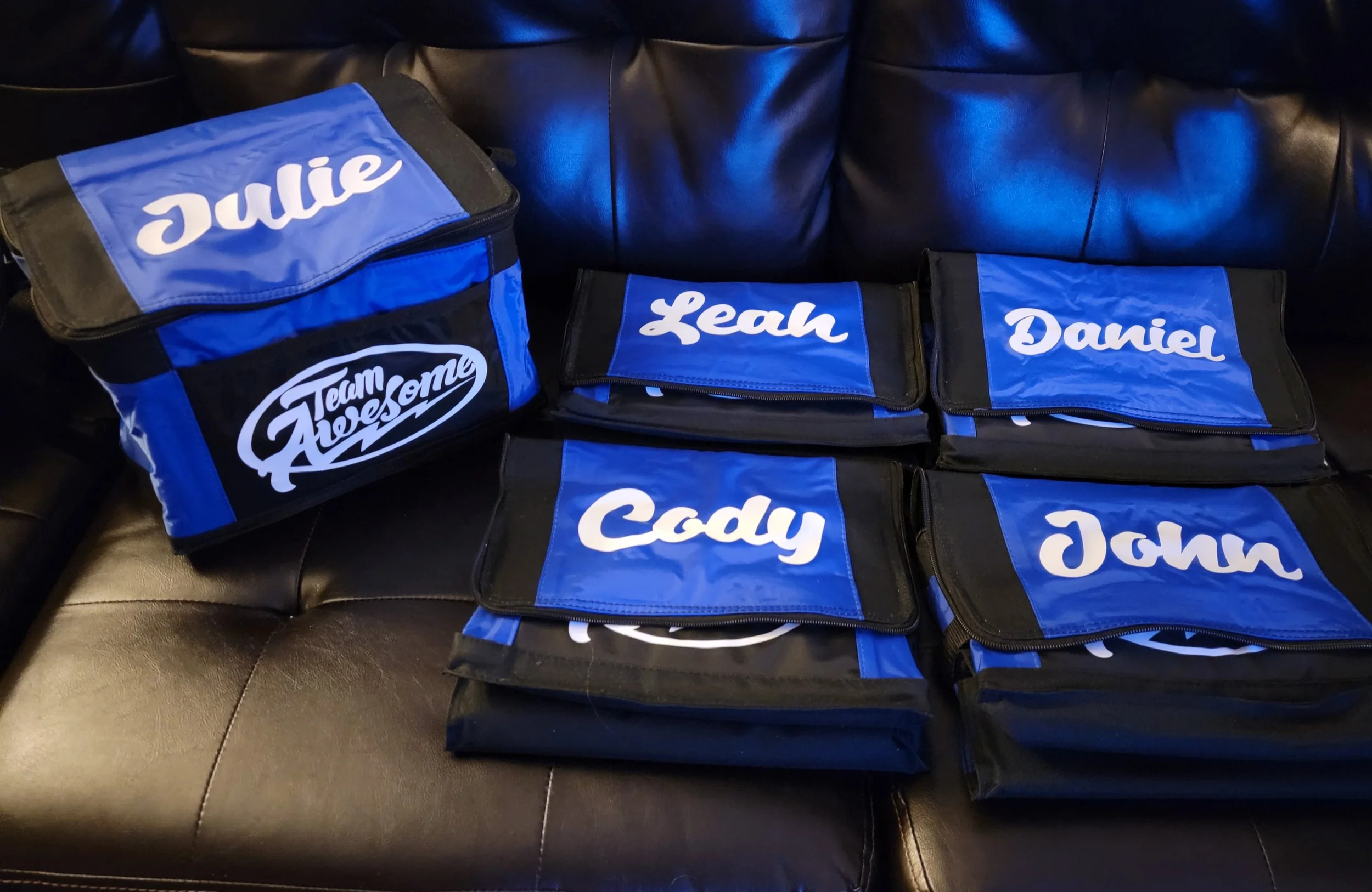 Collection of personalized blue and black bags with white cursive names, some labeled with 'Team Awesome' logo, laid out on a black leather couch.