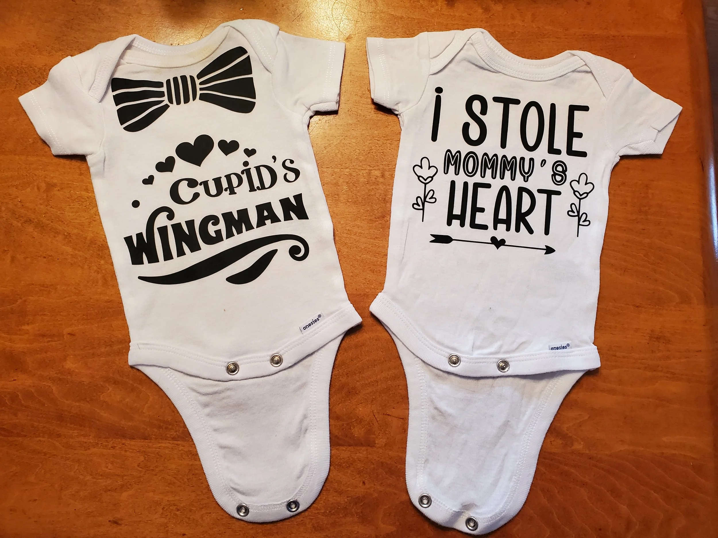 Two white baby bodysuits with black text and graphics. The left bodysuit has a large bow tie graphic and the words "Cupid's Wingman" with hearts. The right bodysuit has the phrase "I stole mommy's heart" with small flower illustrations and an arrow g