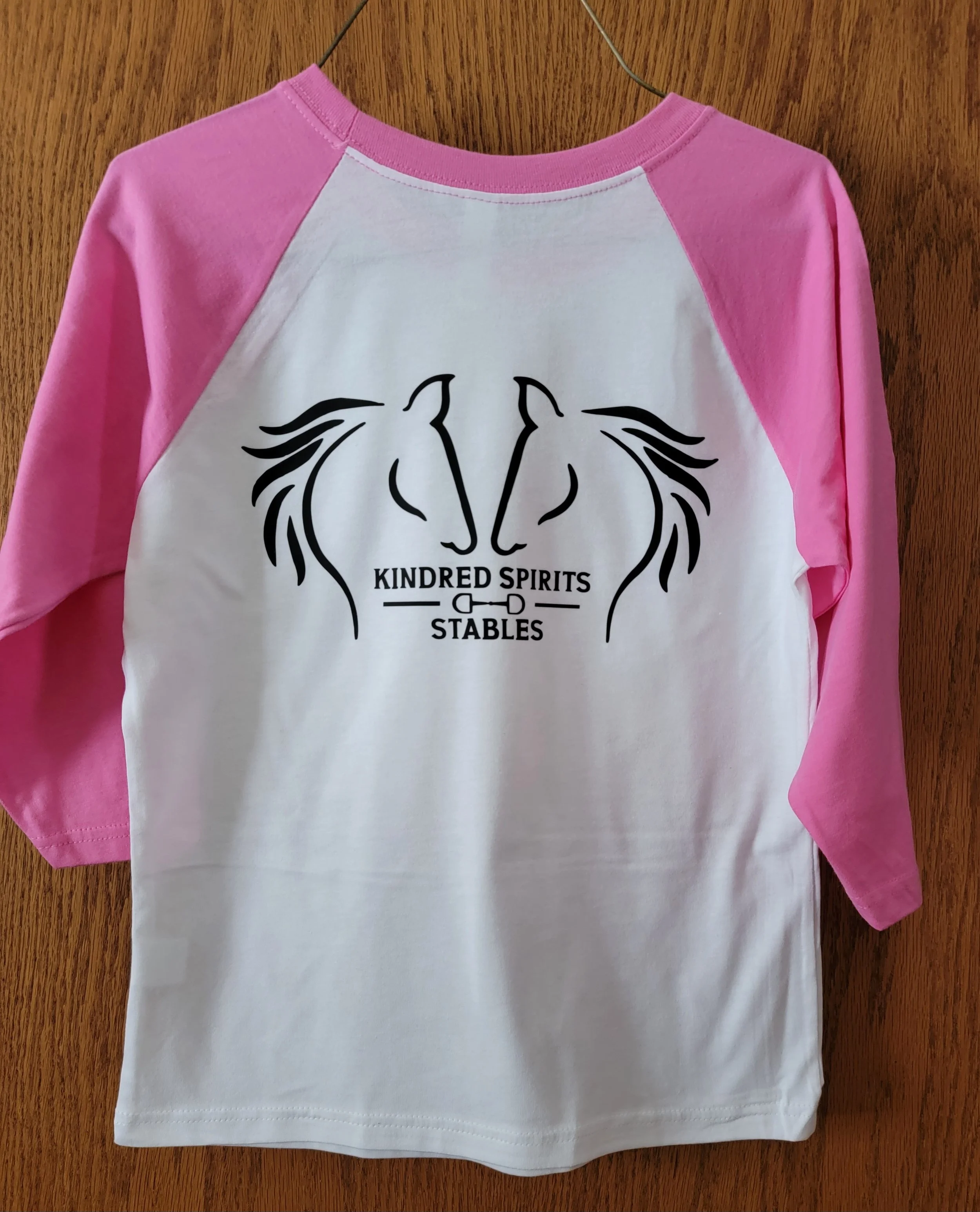 Pink and white baseball-style shirt with a black logo of two stylized horses' heads facing each other and the words "Kindred Spirits Stables" underneath.