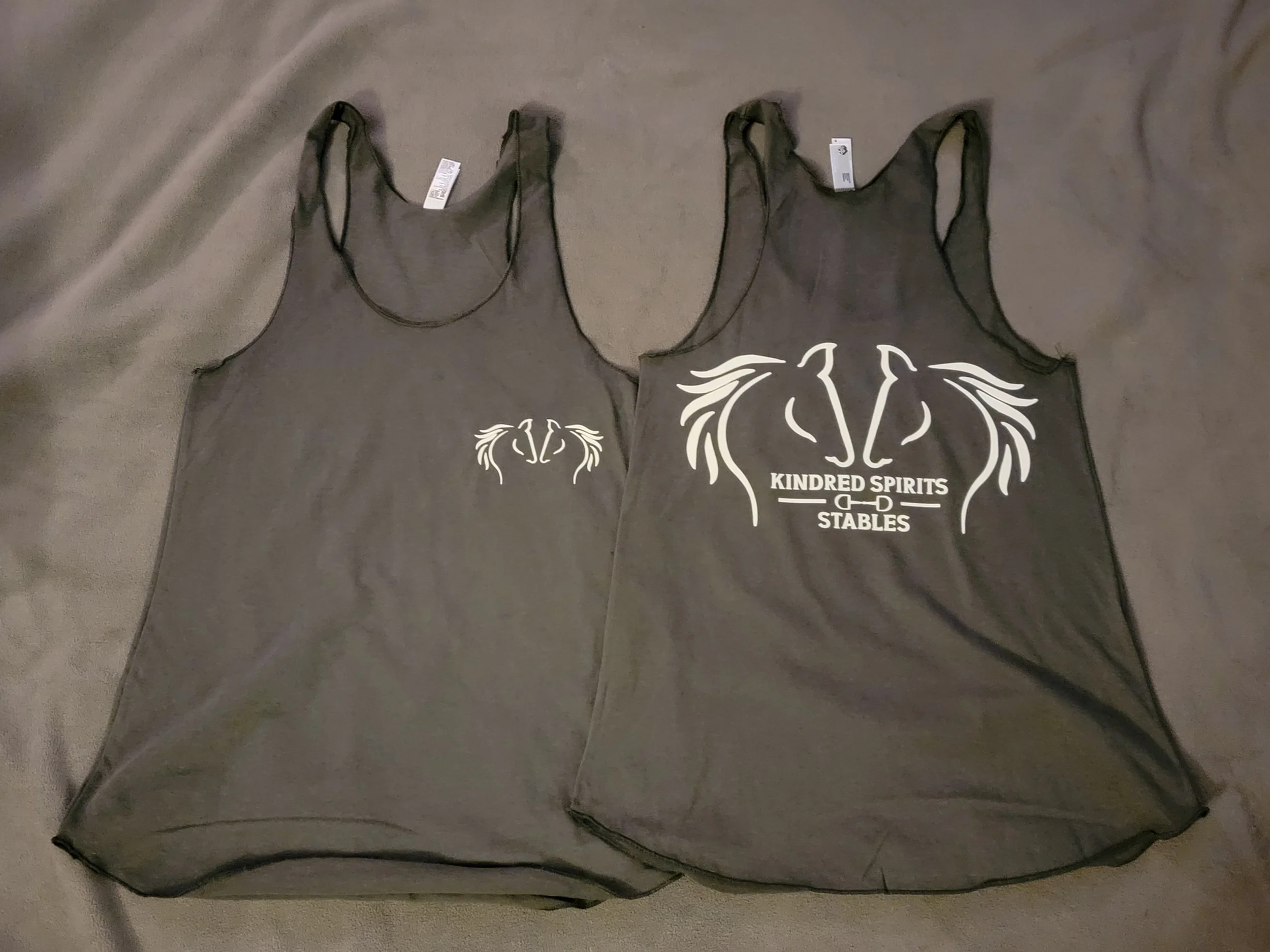 Two black tank tops lying on a surface. The tank top on the right has a white graphic design with stylized horse heads and the text 'KINDRED SPIRITS STABLES'.