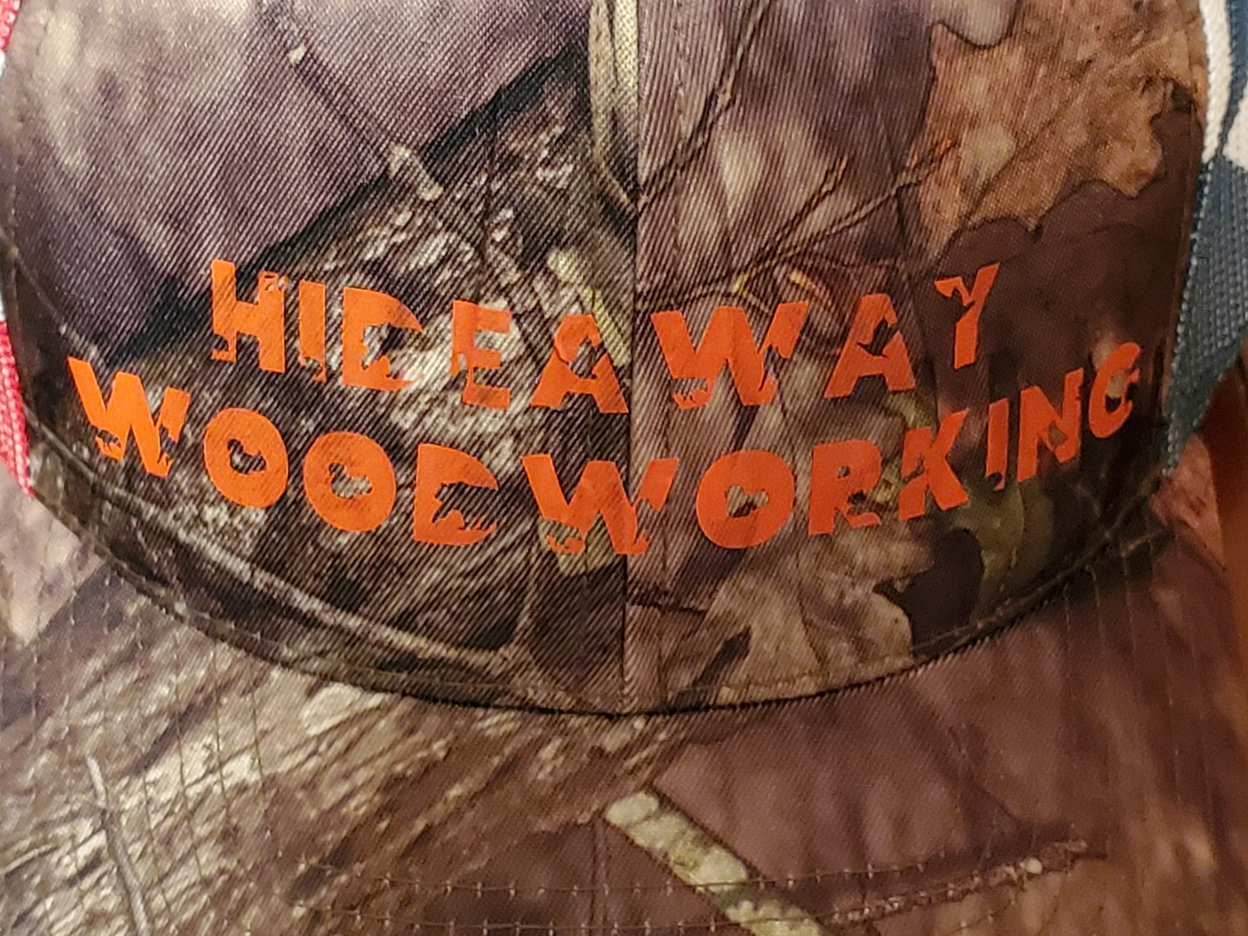 Close-up of a camouflage cap with orange text that reads 'HIDEAWAY WOODWORKING'.
