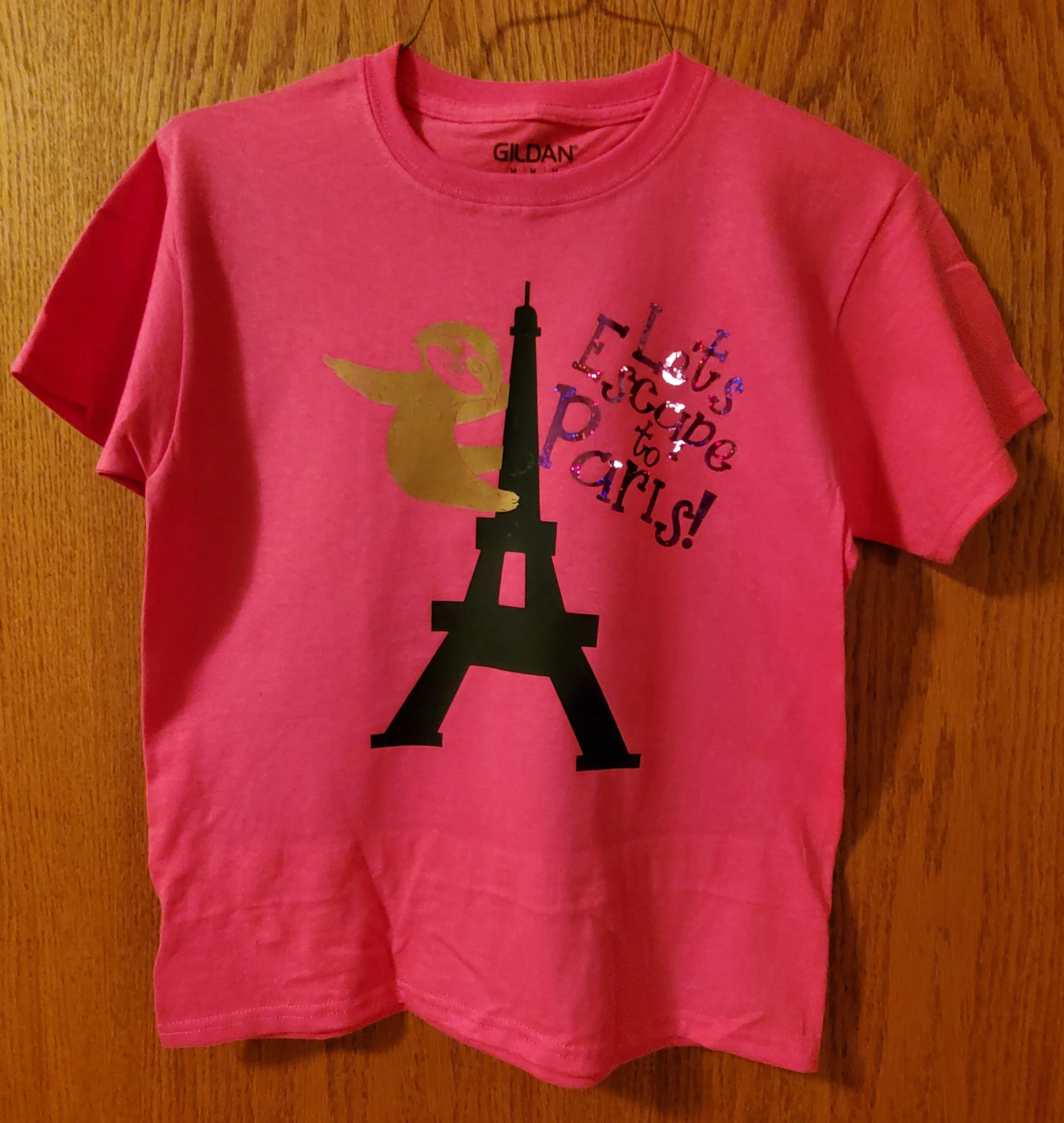 Pink T-shirt with a graphic of the Eiffel Tower, a cartoon bunny holding a finger to its lips, and the text "Let's be Jueparis!" in colorful letters.