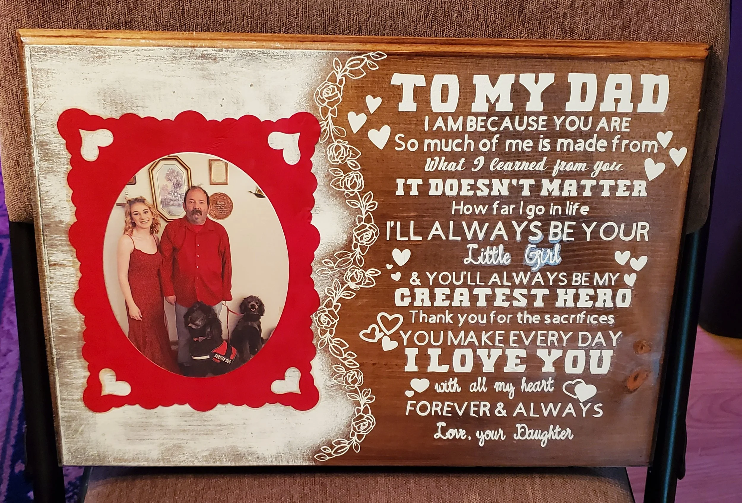 A framed display with a photo of a daughter and her father standing with two service dogs, surrounded by a decorative red border and a heartfelt message to her dad.