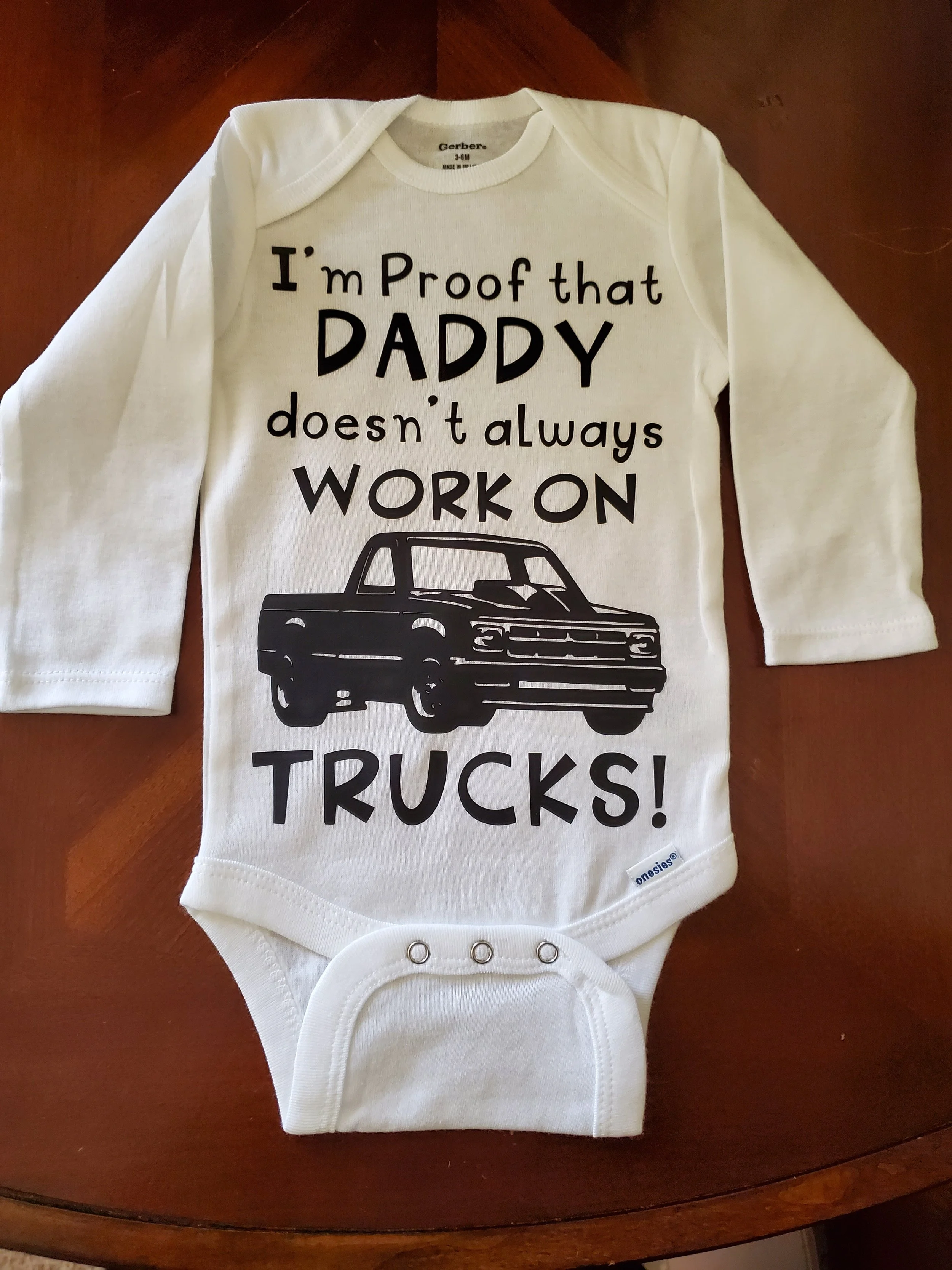 White baby onesie with black text and a truck illustration. The text reads, "I'm Proof that DADDY doesn't always WORK ON TRUCKS!".
