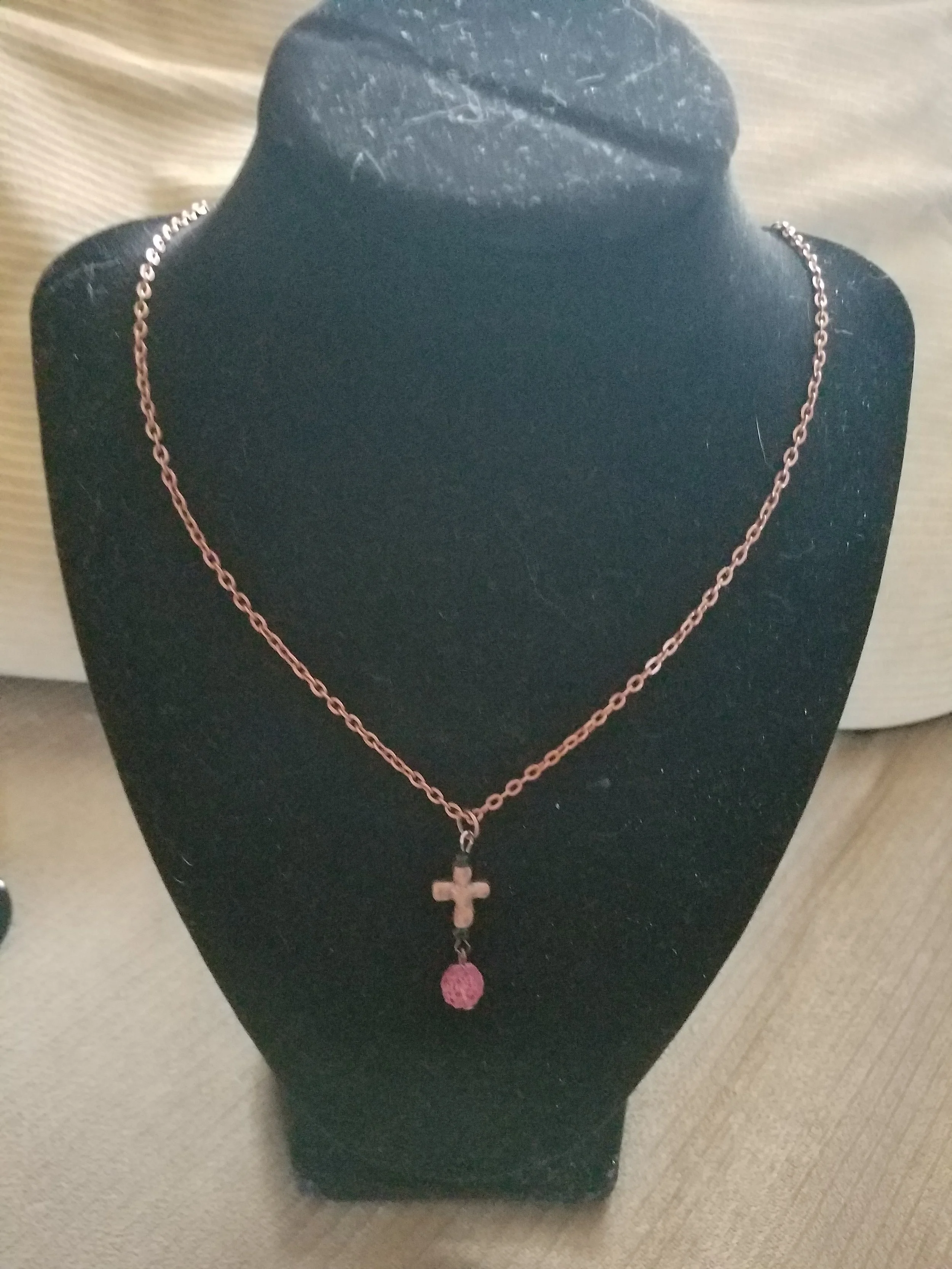A necklace with a cross pendant and a pink bead hangs on a black necklace display bust.