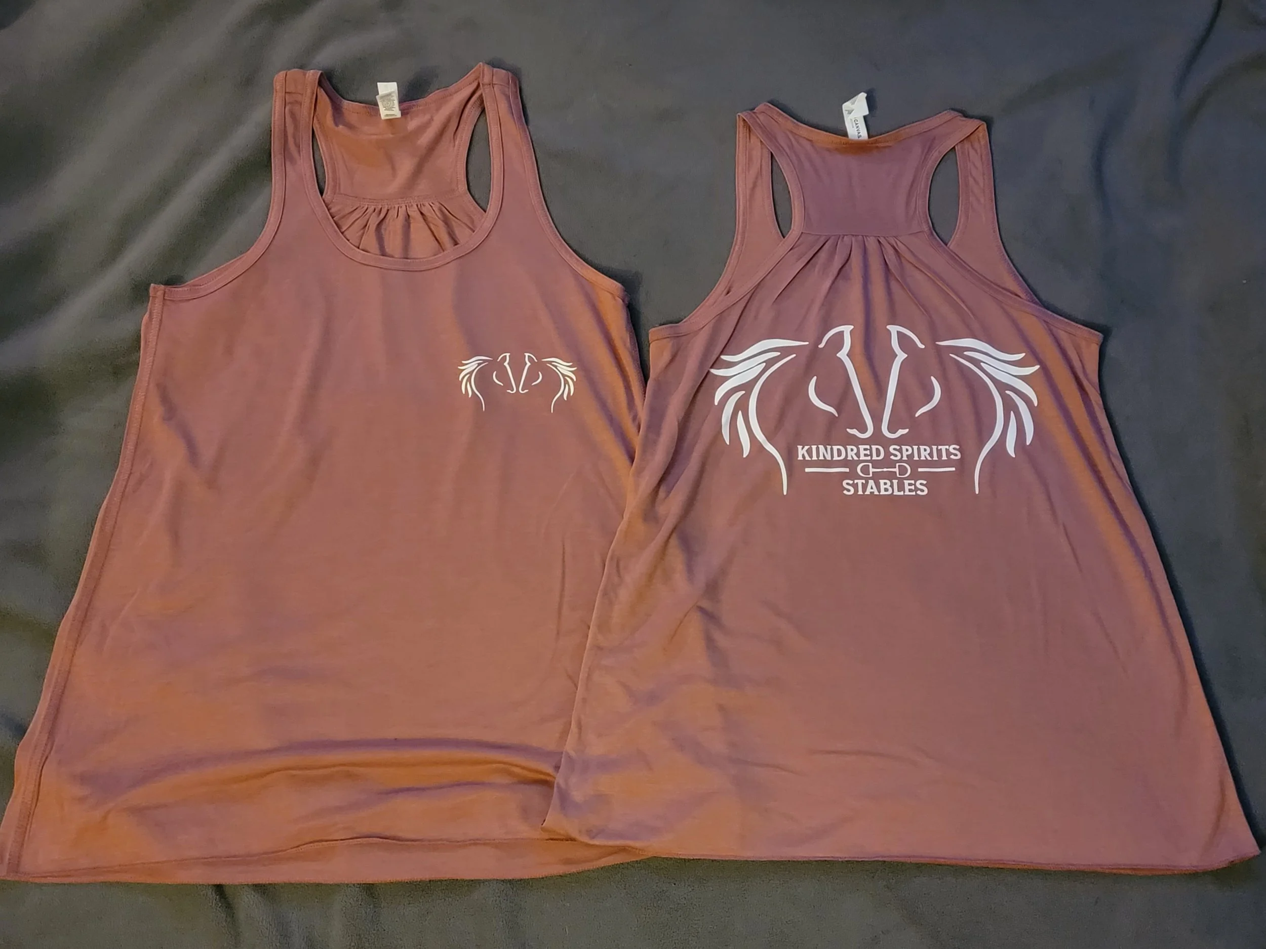 Two matching brown tank tops with a horse-themed logo. The smaller tank top has a small white logo on the left chest area, and the larger tank top has a larger logo on the front with the words "KINDRED SPIRITS STABLES" and a stylized horse head illus