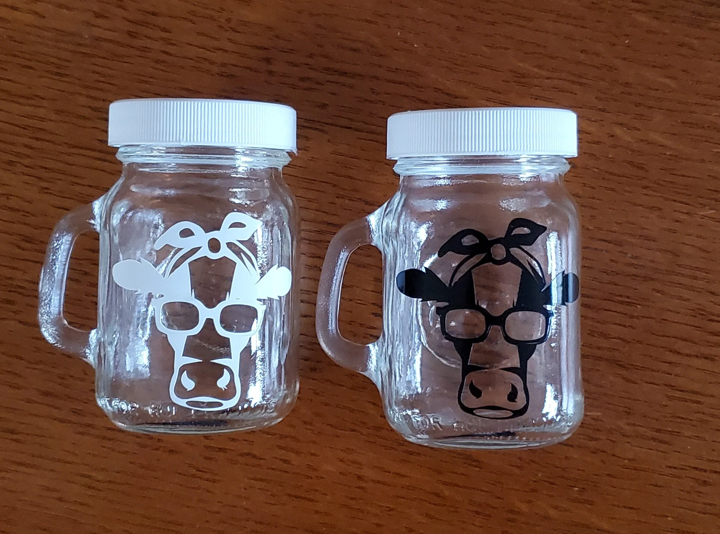 Two small glass jars with white lids and cow-themed designs on the front, placed on a wooden surface. One features a white cow head with glasses and a bow; the other has a black cow head with glasses and a bow.