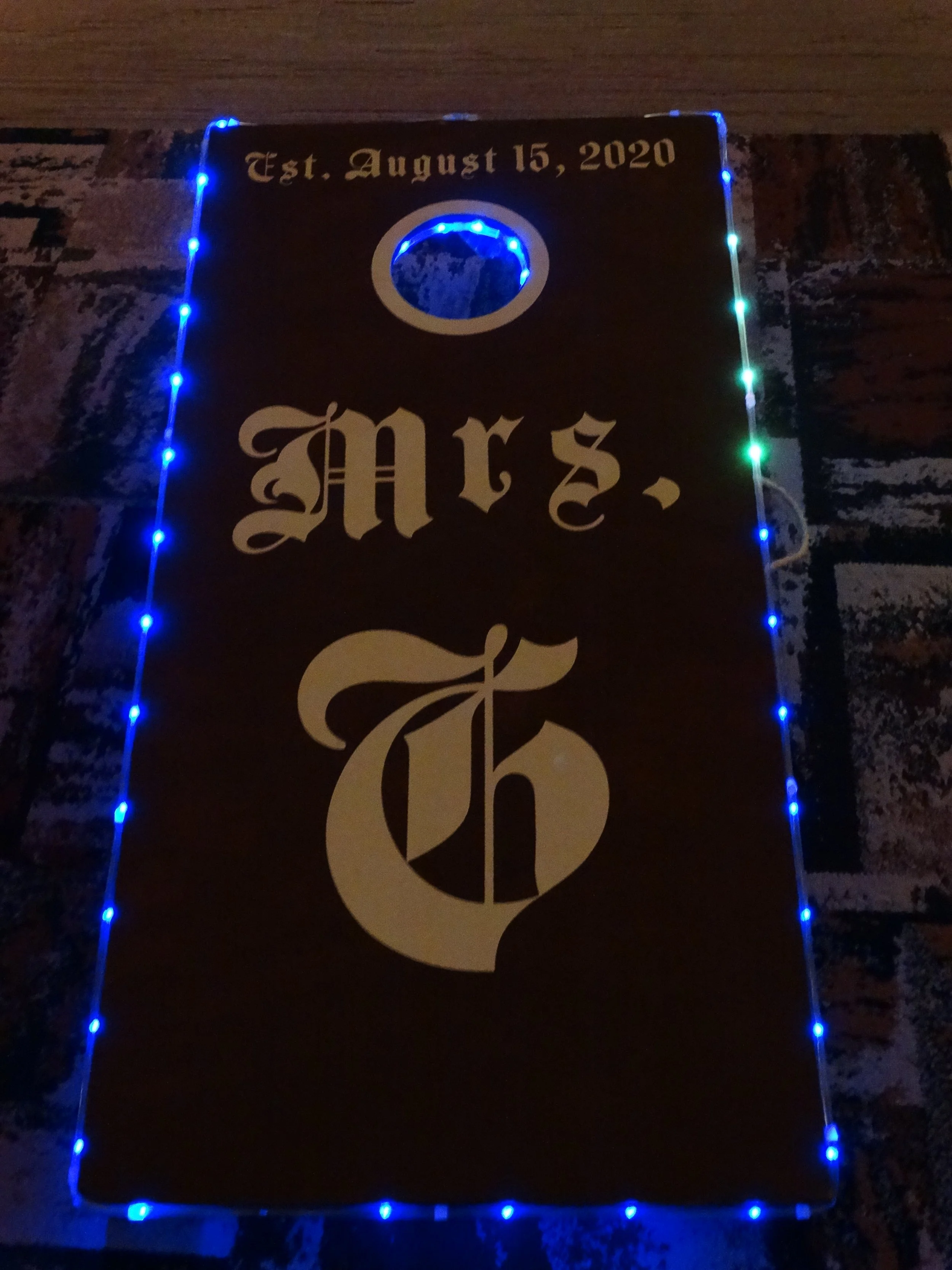 A black banner with white gothic text reads 'Est. August 15, 2020' at the top, surrounded by blue and some green string lights. The banner has a circular hole near the top center and a large stylized letter 'C' near the bottom.