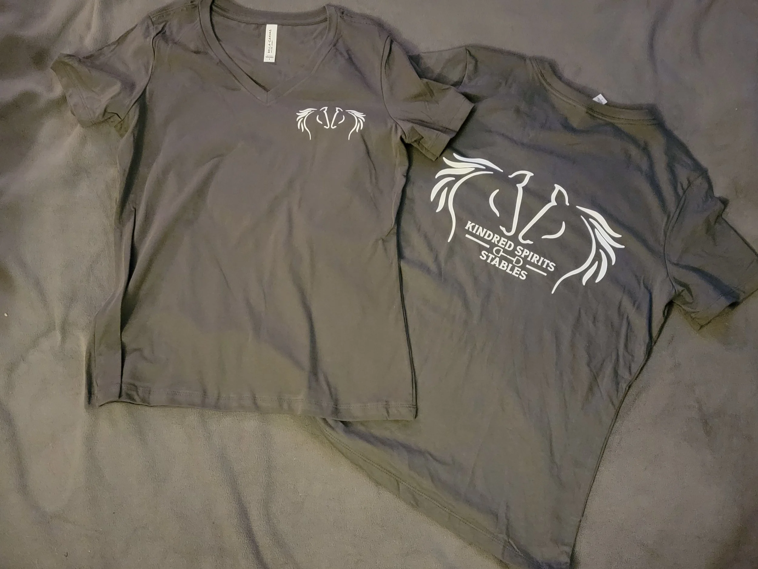 Gray T-shirt with white logo and text reading 'Kindred Spirits Stables' laid flat on a gray surface.