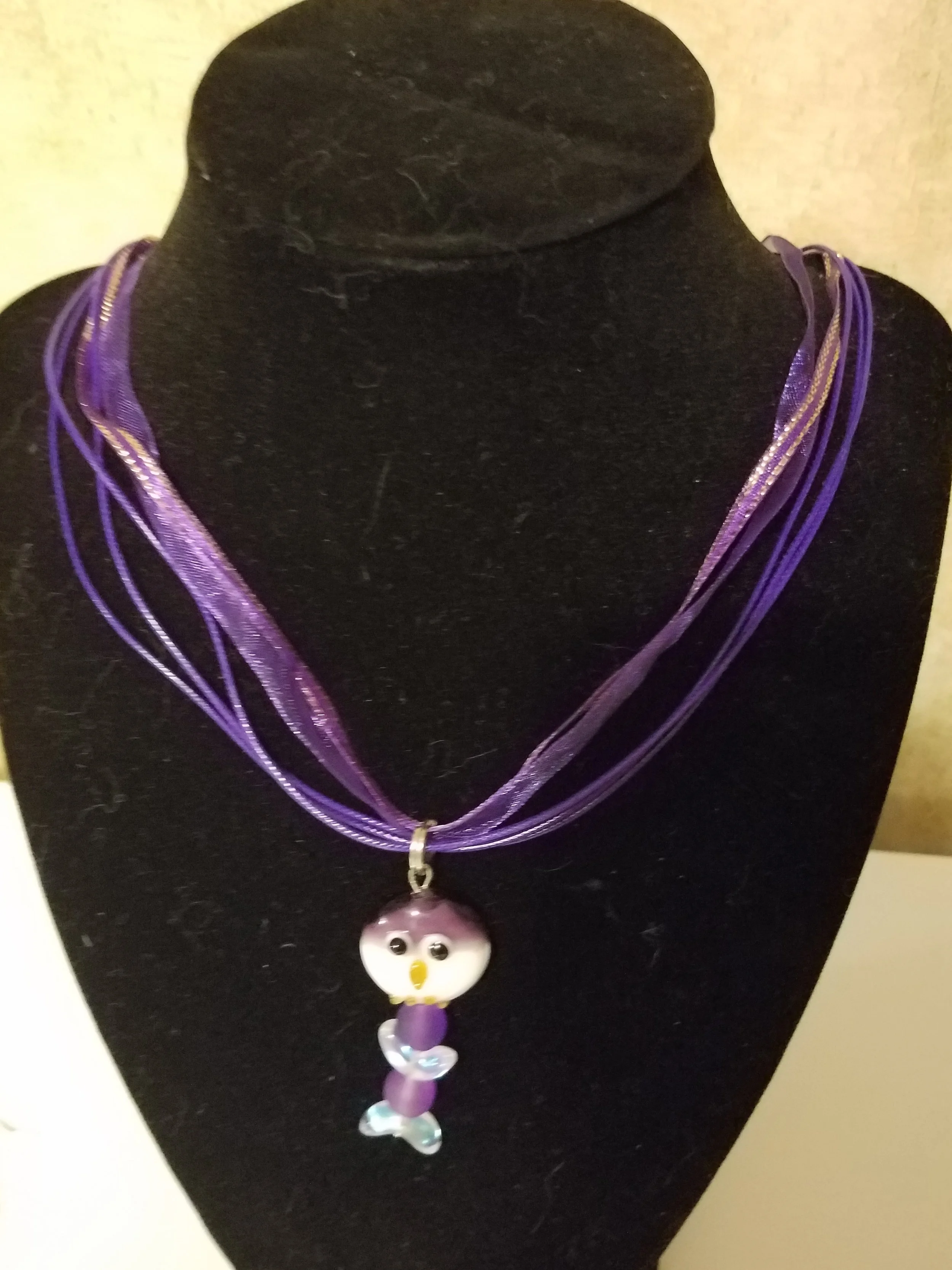 Necklace with purple and lavender multi-strand cords and a puffy owl pendant on a black display bust.