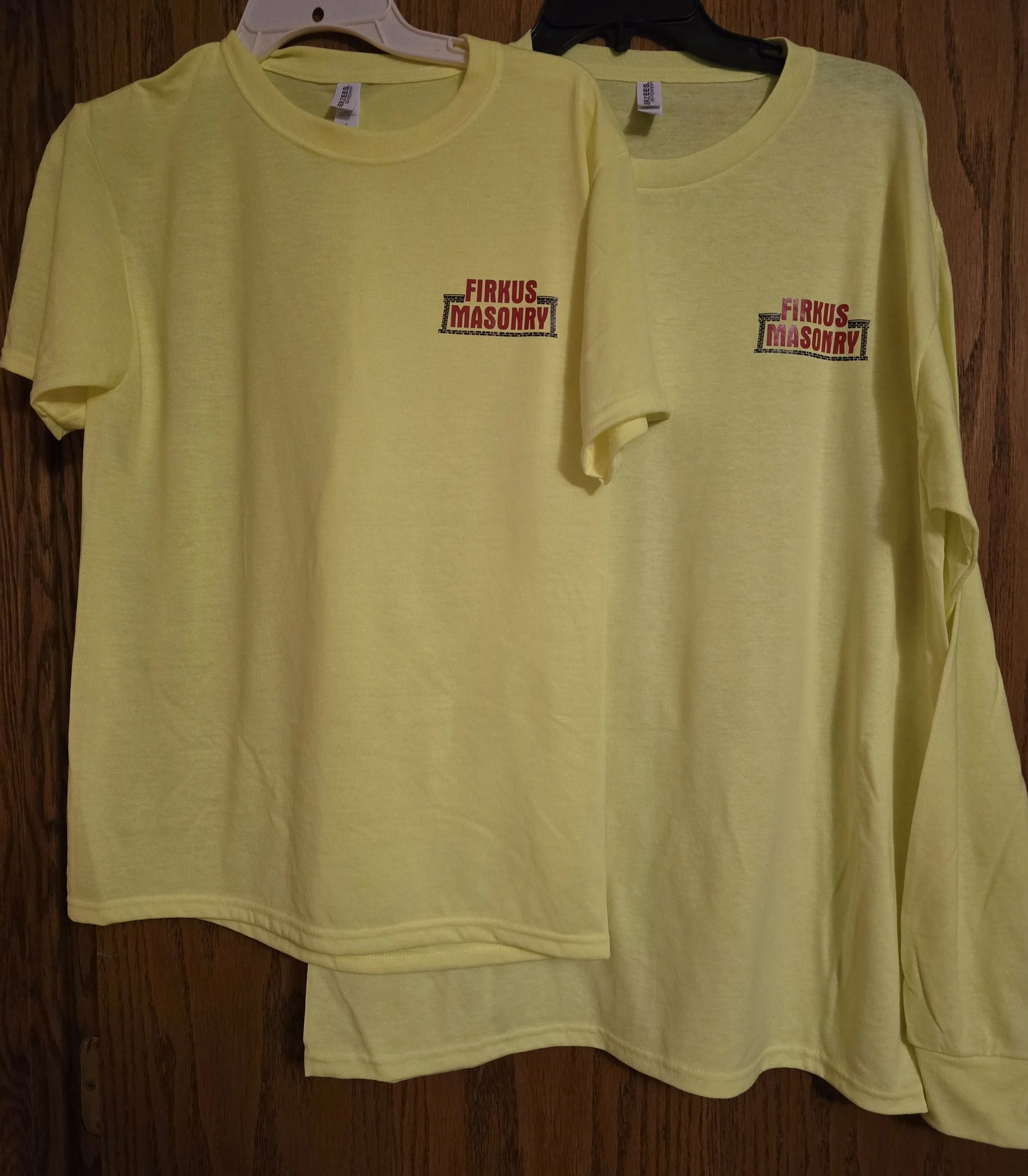 Two yellow T-shirts hanging on hooks on a wooden wall, each with a logo reading "FIRKUS MASONRY" in red and black text.