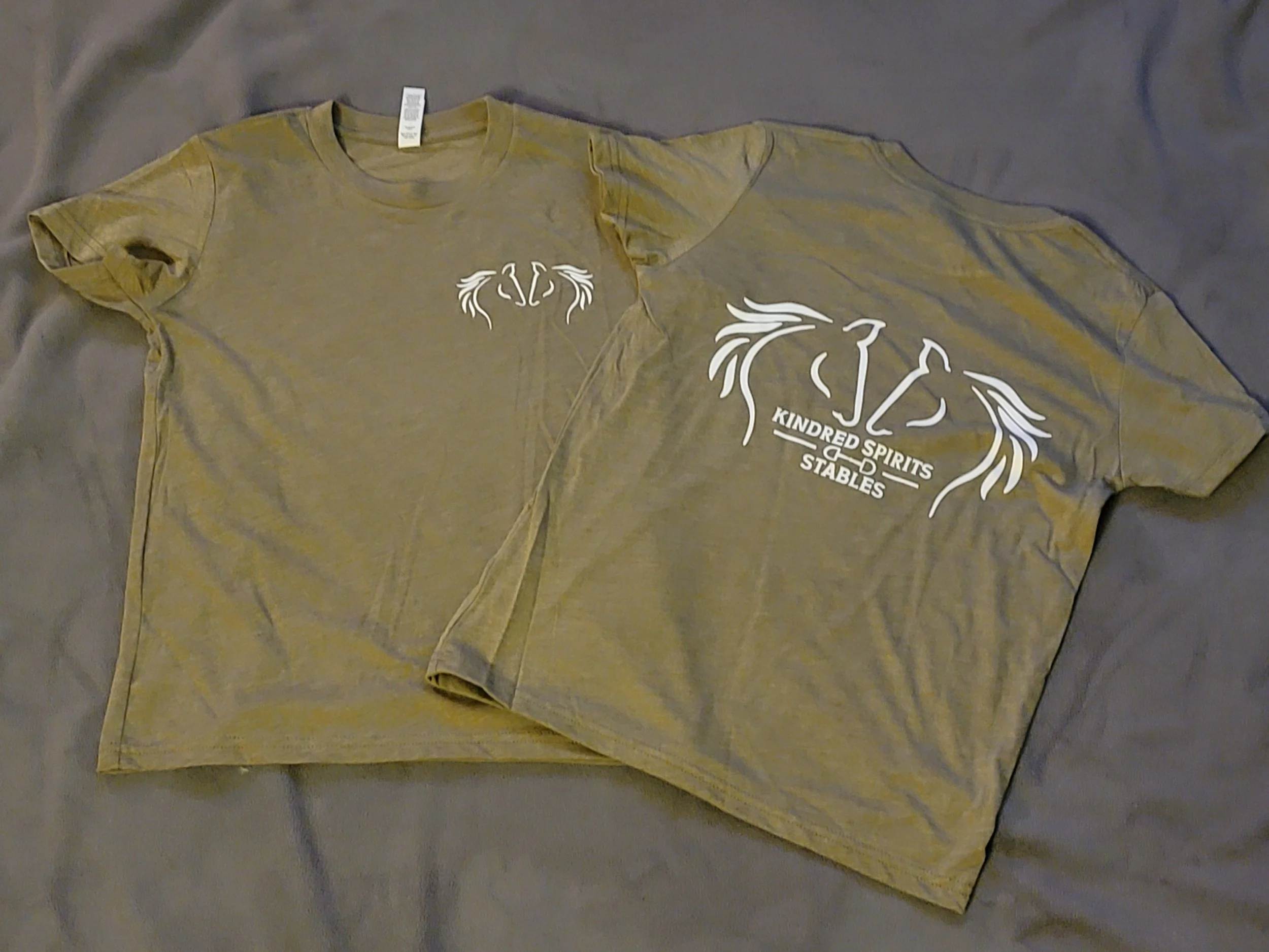 Two beige T-shirts, one with a small design and the other with a larger design with text, are laid flat on a gray fabric surface.