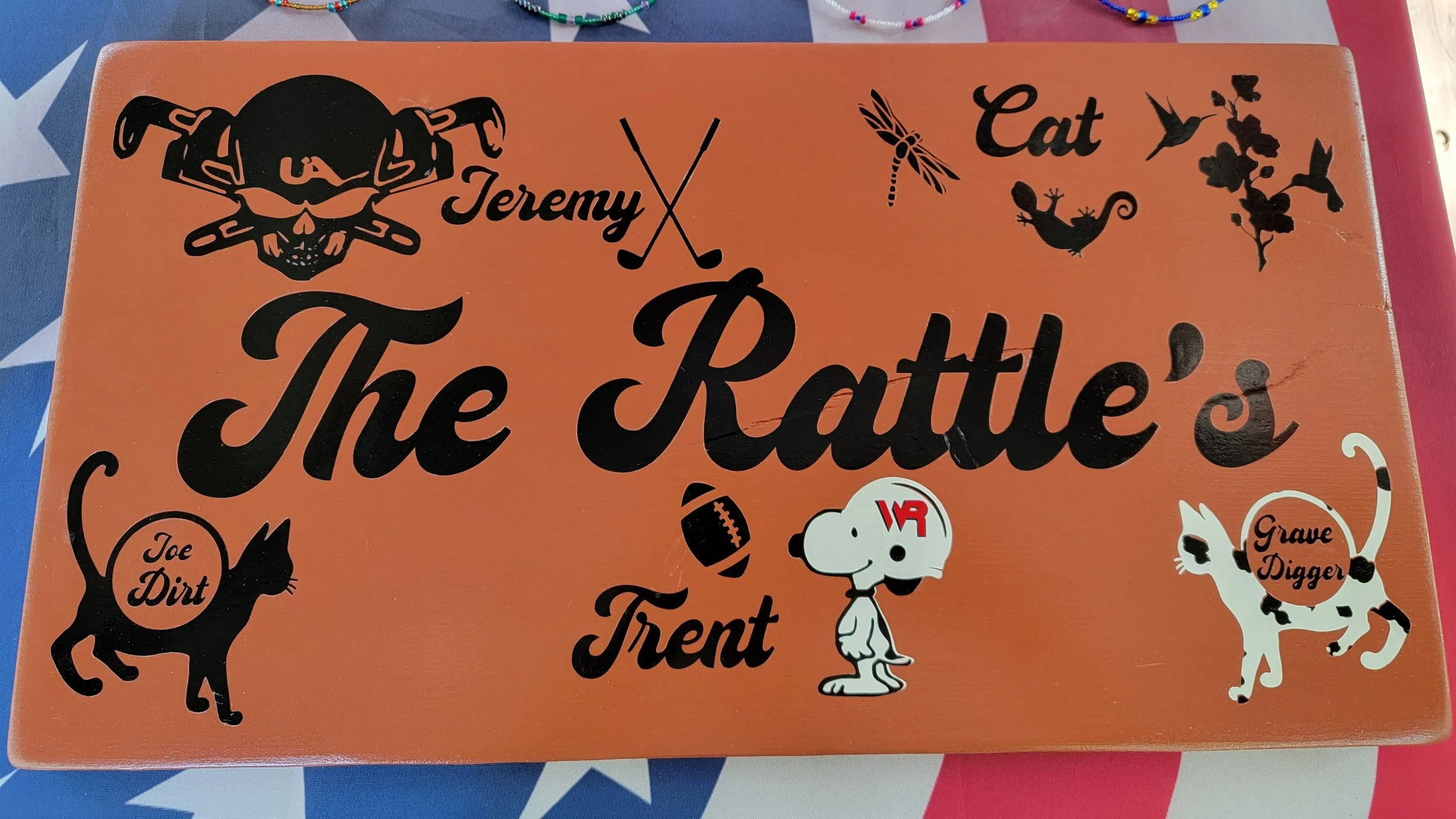 Decorative sign with black illustrations and text on an orange background. It features a skull and crossbones with pigtails, a football, a dog wearing a helmet, a cat, a dragonfly, hummingbirds, and a plant. The text includes "Jeremy," "Trent," "Joe 