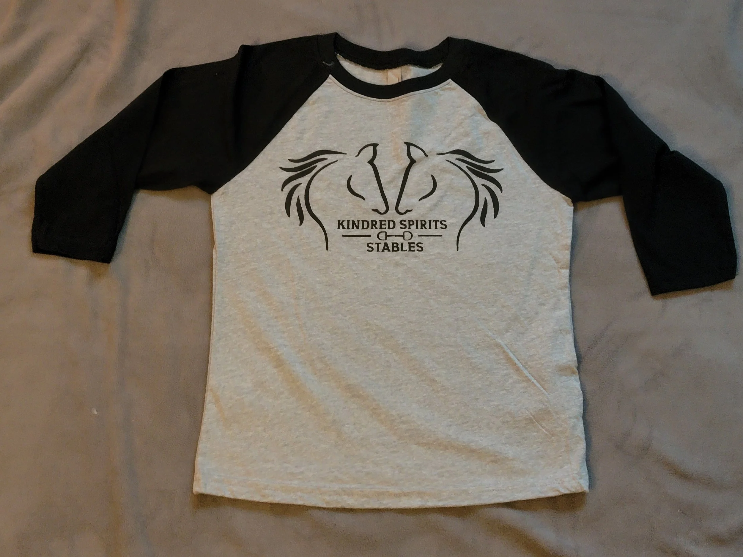 A raglan style baseball T-shirt with black sleeves and a gray body, featuring a graphic of two horses' heads and the text 'KINDRED SPIRITS STABLES'.