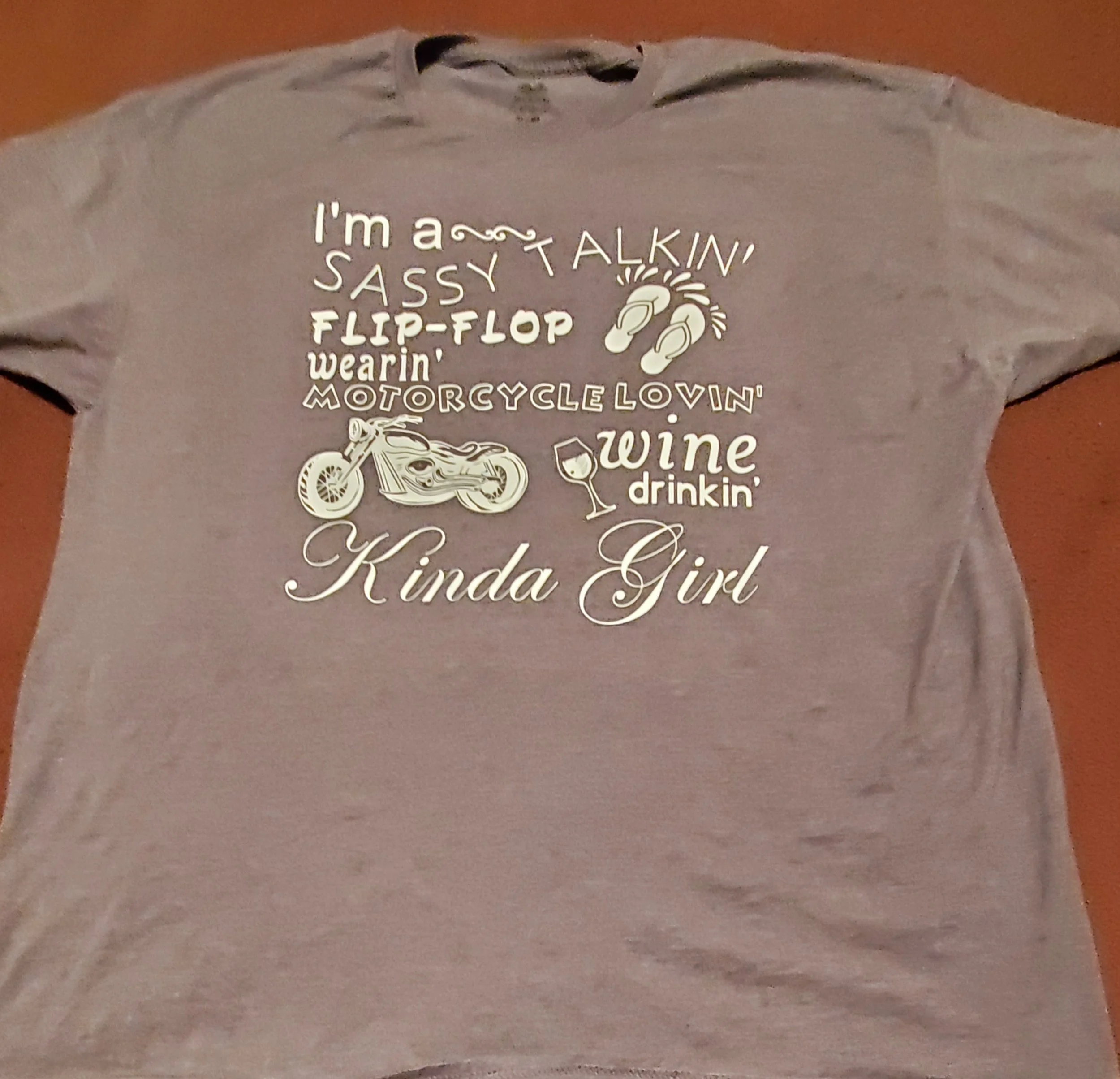Gray t-shirt with white text and illustrations that say, "I'm a sassy talkin', flip-flop wearin', motorcycle lovin' kinda girl" with images of flip-flops, a motorcycle, and a wine glass.