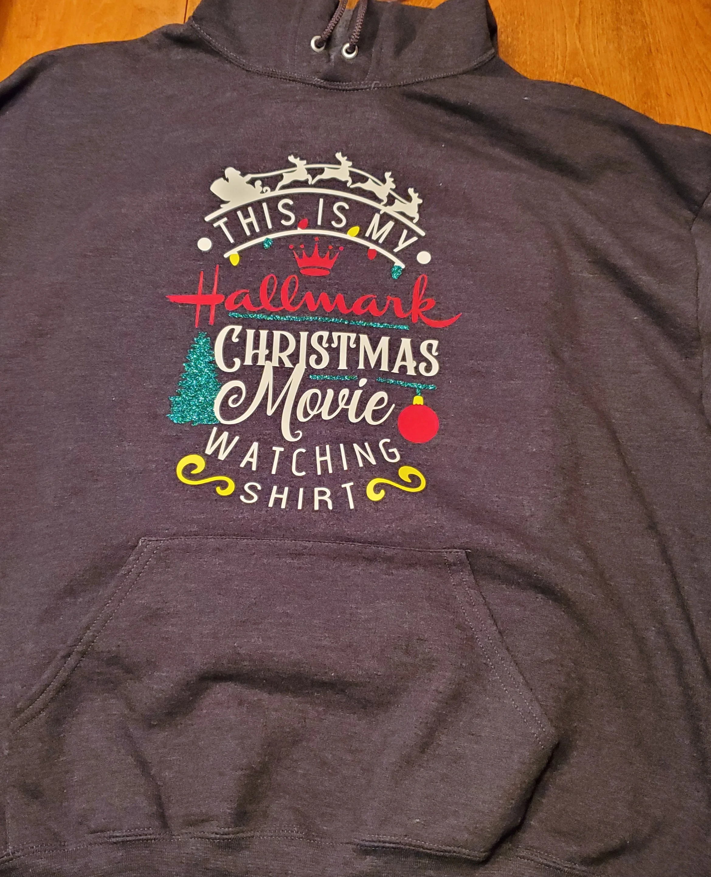 Black hoodie with Christmas-themed text and images, including a Santa silhouette on a sleigh, a Christmas tree, a red ornament, and festive decorative elements.