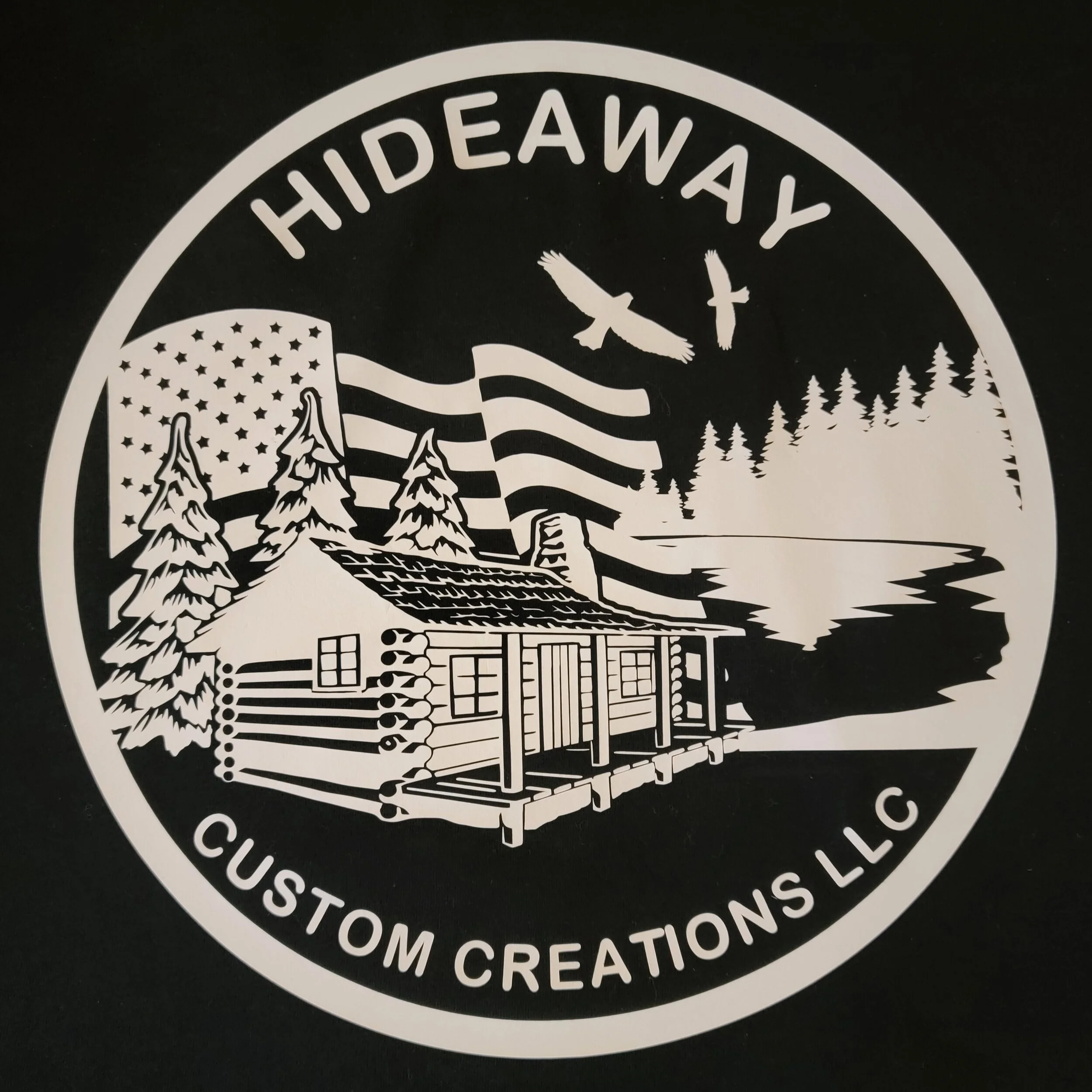 Black and white circular logo for Hideaway Custom Creations LLC featuring a cabin, pine trees, hills, a lake, American flag, and two flying birds.
