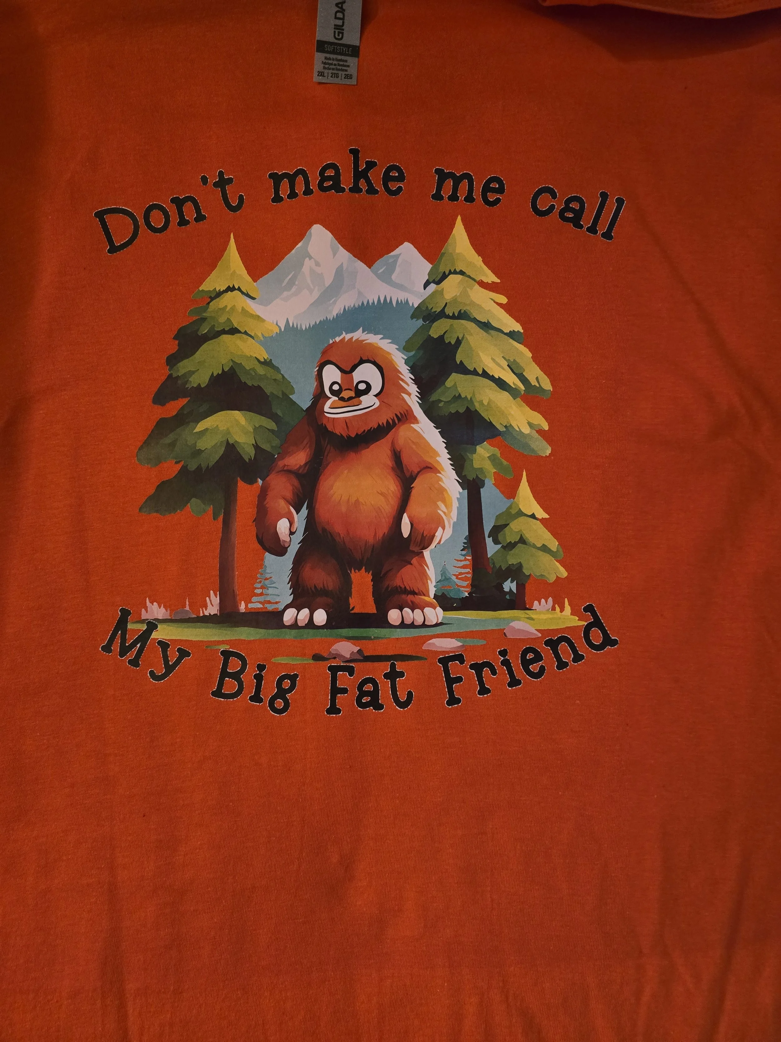 Orange T-shirt with a cartoon illustration of a Bigfoot creature in front of trees and mountains, with the text 'Don't make me call My Big Fat Friend'.