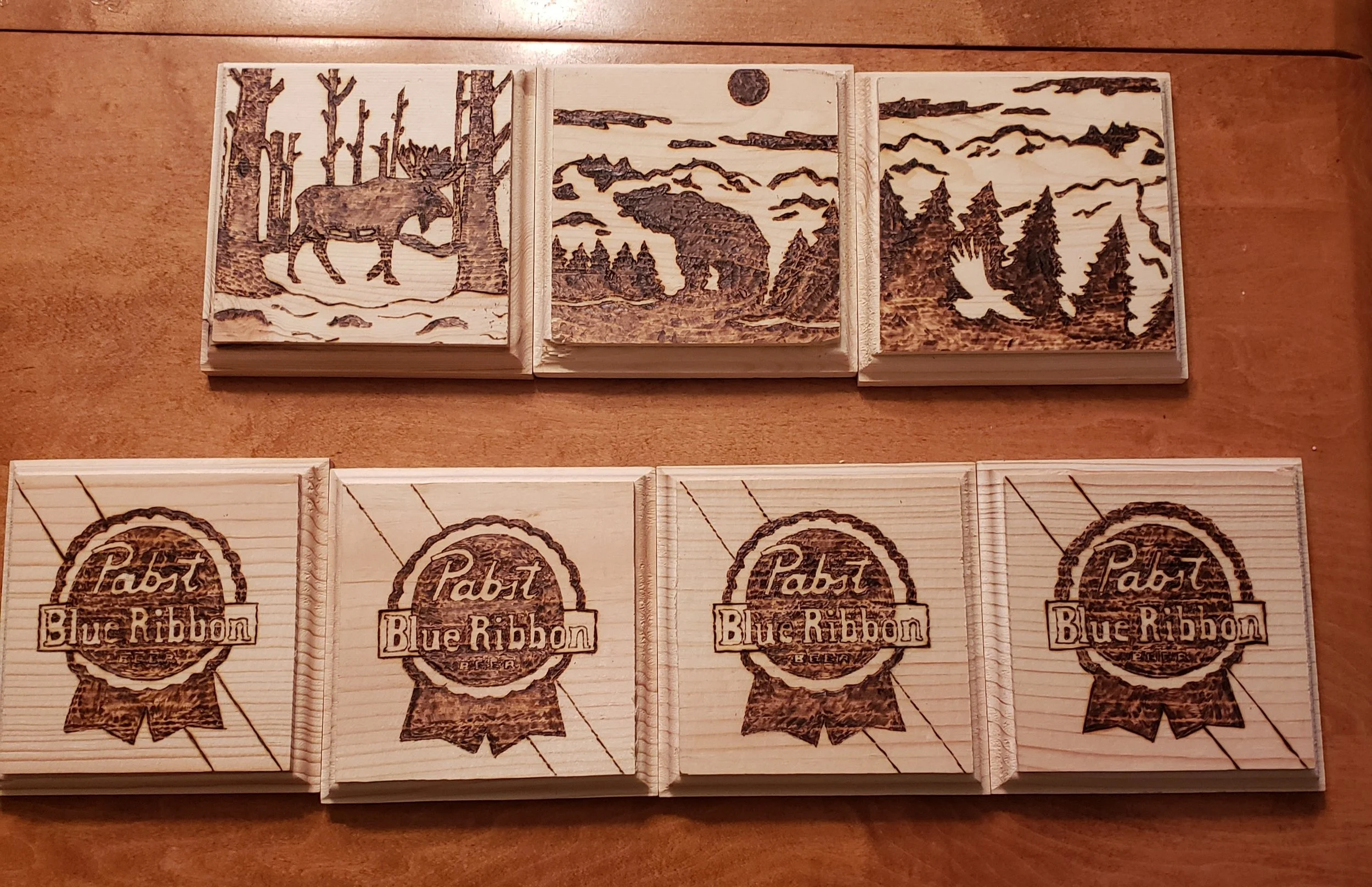 Sets of wooden blocks with engraved images. The top row features forest scenes: a moose among trees, a bear under a full moon, and a forest with a bird flying among trees. The bottom row displays branded blocks with the Pabst Blue Ribbon logo.